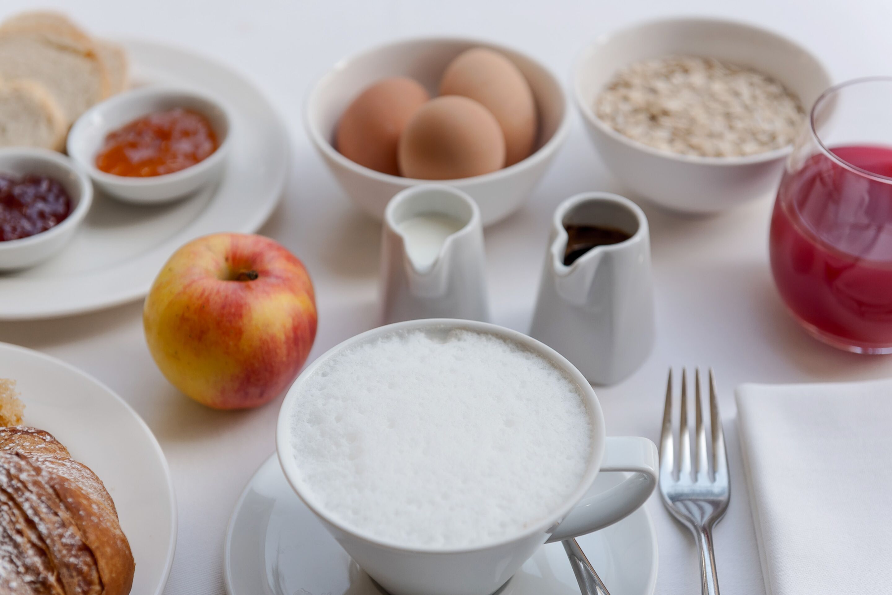 A breakfast setting with a plate of toast, a bowl of eggs, a bowl of cereal, a jug of milk, a glass of juice, a cup of coffee, and a slice of cake.