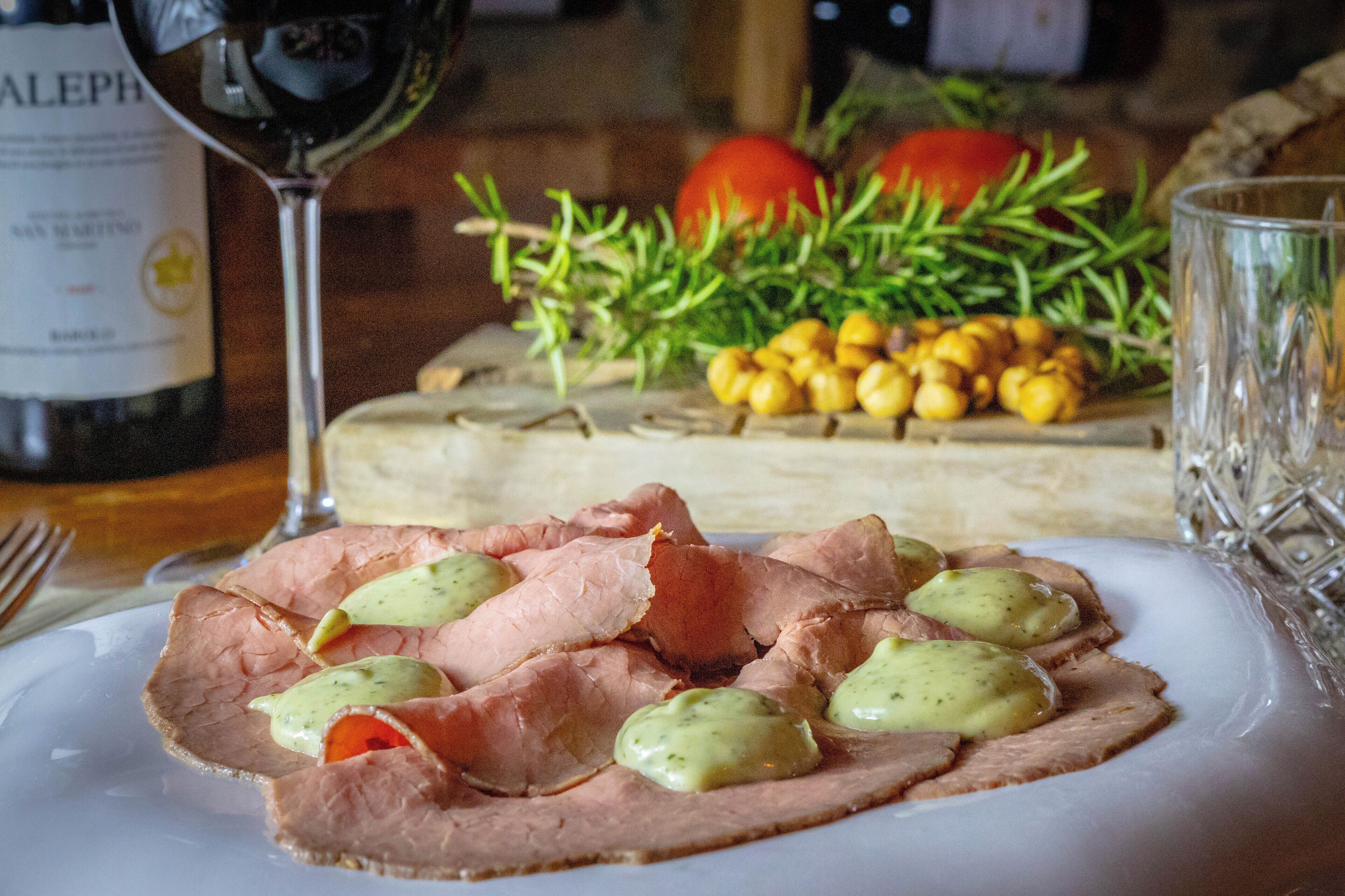 Un piatto di prosciutto a fette con salsa cremosa, un bicchiere di vino rosso e un tagliere di legno con verdure ed erbe aromatiche sullo sfondo.