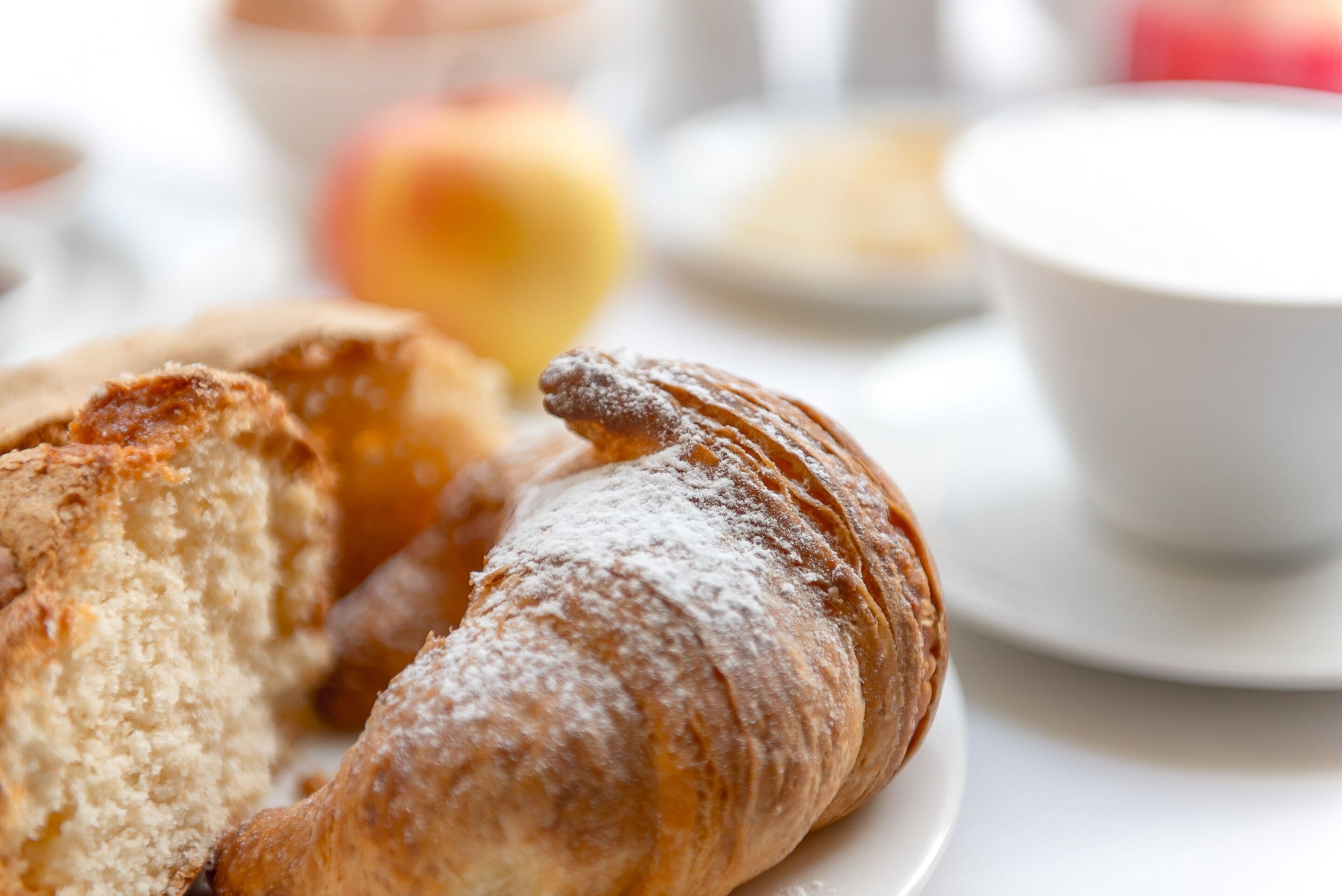A croissant on a plate with a blurred background of a cup and a fruit.