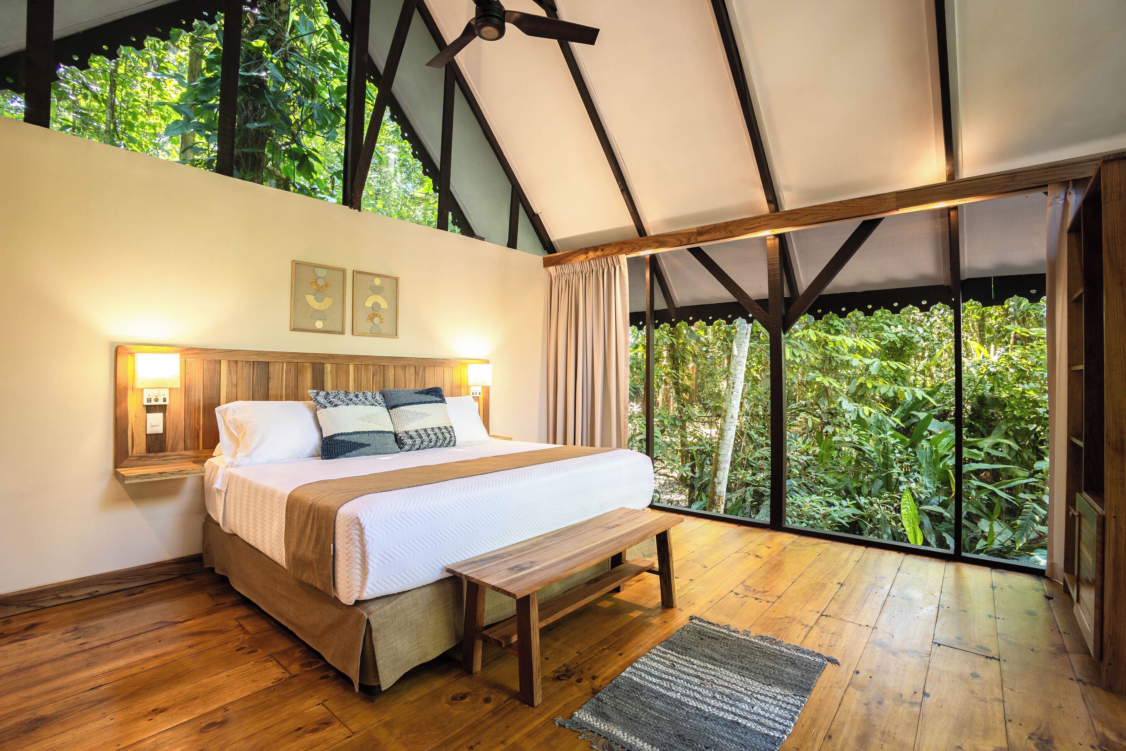 Quarto com uma cama grande, cabeceira de madeira, abajures e vista para o verde através de grandes janelas.