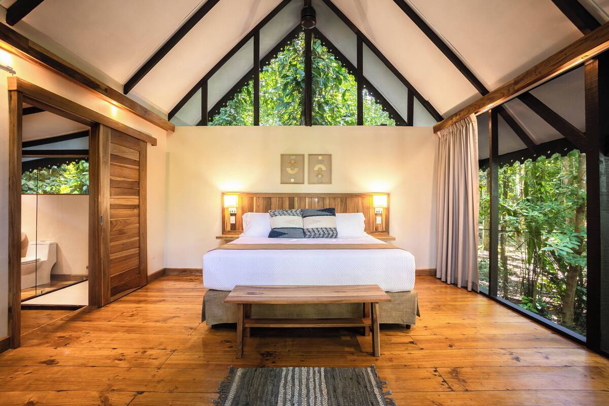 A bedroom with a large bed, wooden flooring, a wooden bench, and a view of greenery through a window.