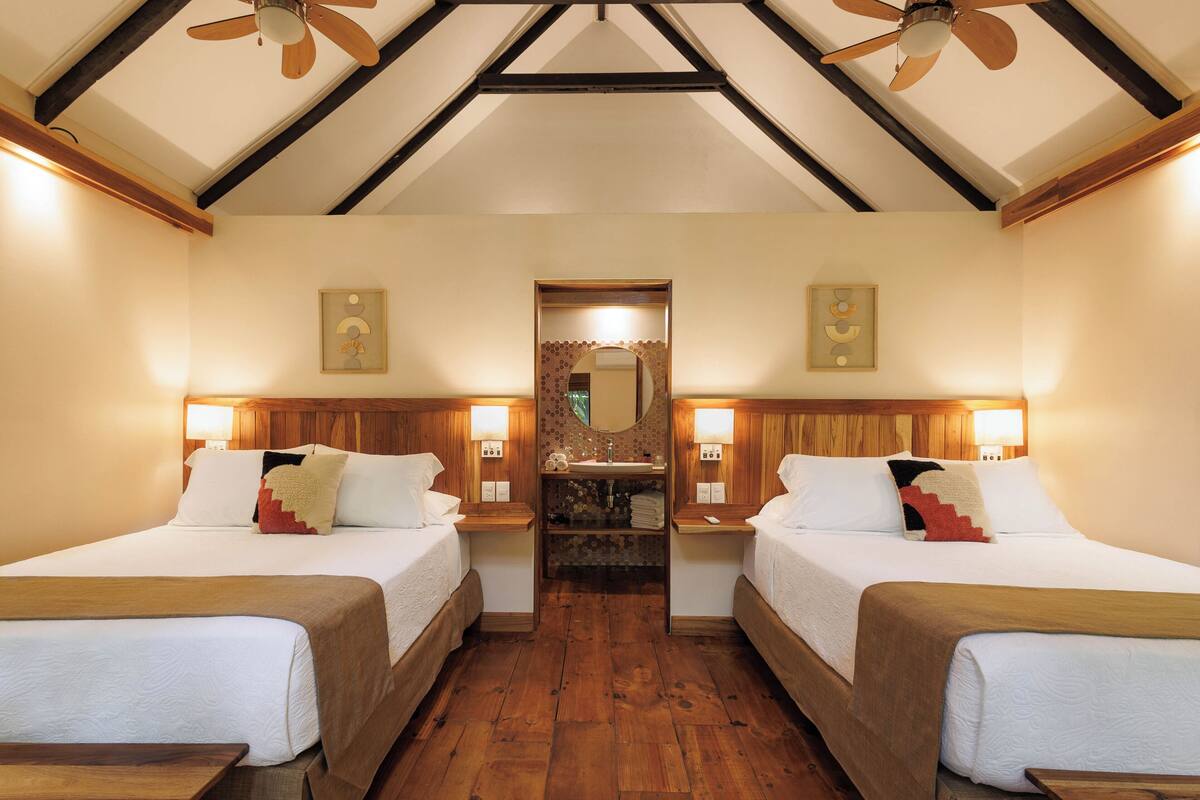 A hotel room with two beds, wooden floors, a ceiling fan, and a mirror above the sink.