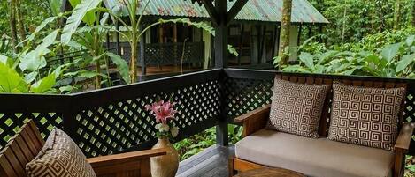 Terraza de madera al aire libre con un área de estar cubierta, que incluye un sofá y sillas, rodeada de abundante vegetación.