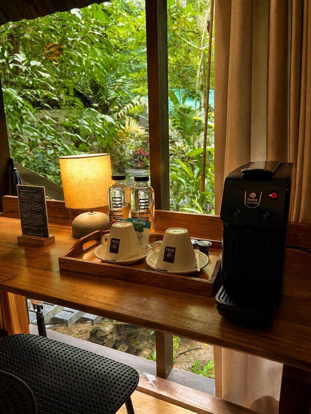 Uma mesa de madeira com uma cafeteira, duas xícaras, um tabuleiro e um abajur.