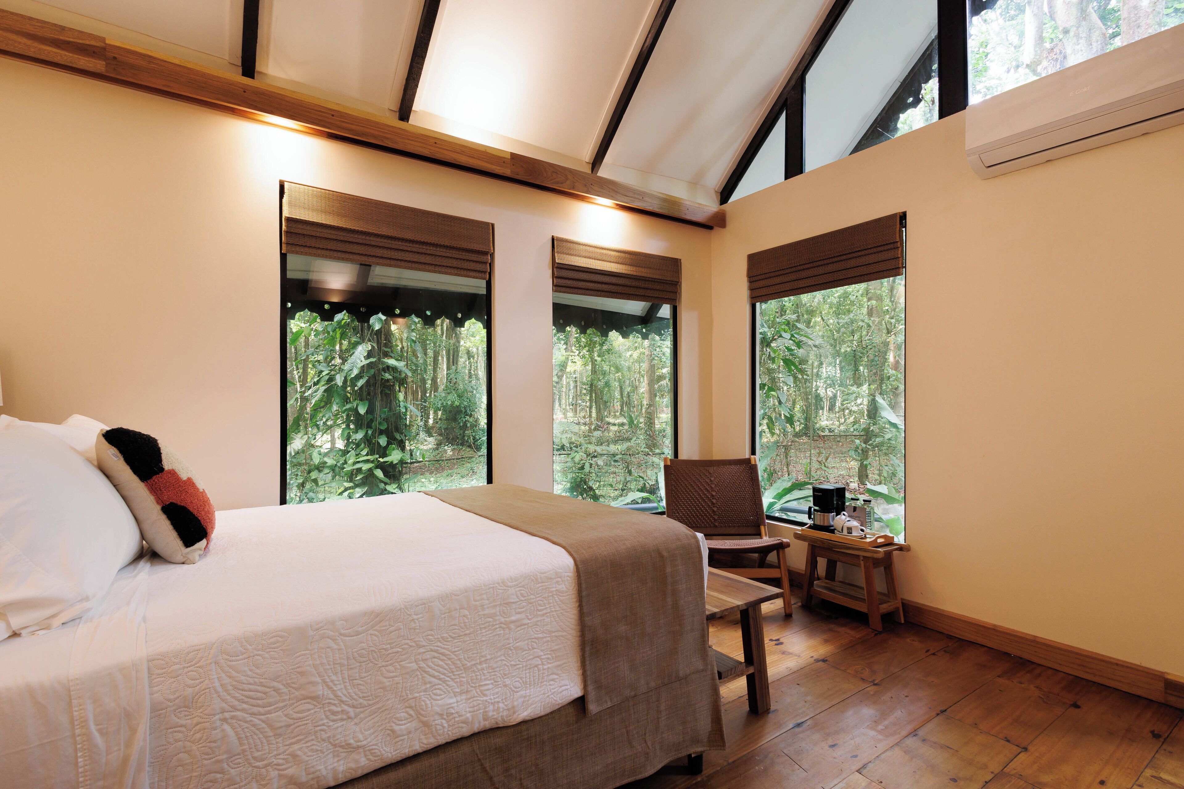 A bedroom with a large bed, a chair, and a small table. There are large windows with blinds, and a ceiling fan is visible.