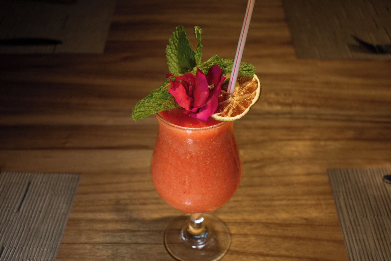 A glass of red beverage with a rose, mint, and orange slice garnish on a wooden table.