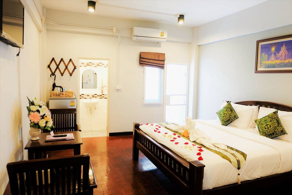 A hotel room with a wooden bed, a small table with a vase of flowers, a mirror, and a window with a view of the outside.