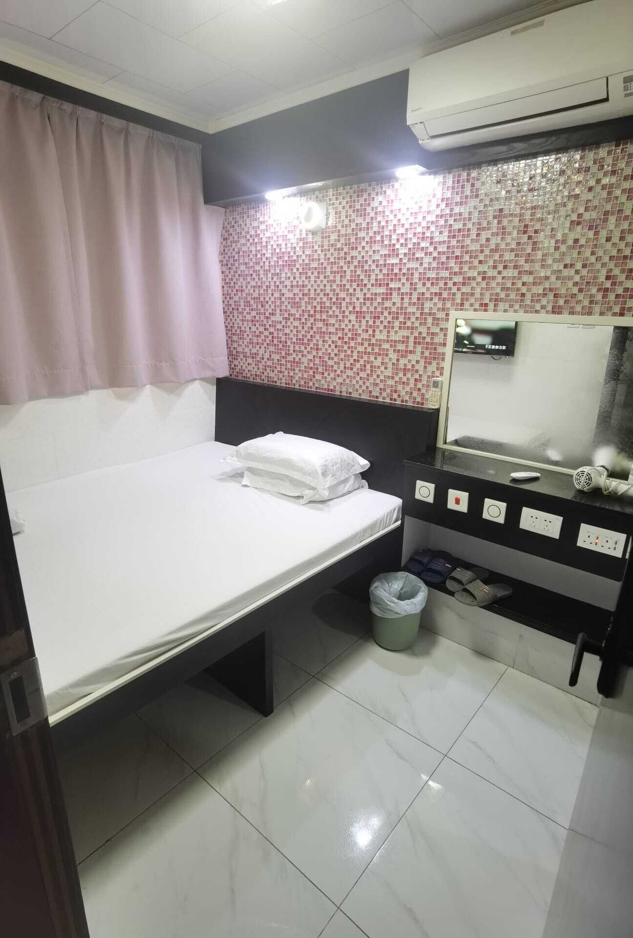 A hotel room with a bed, a small trash bin, a mirror, and a wall-mounted air conditioner.
