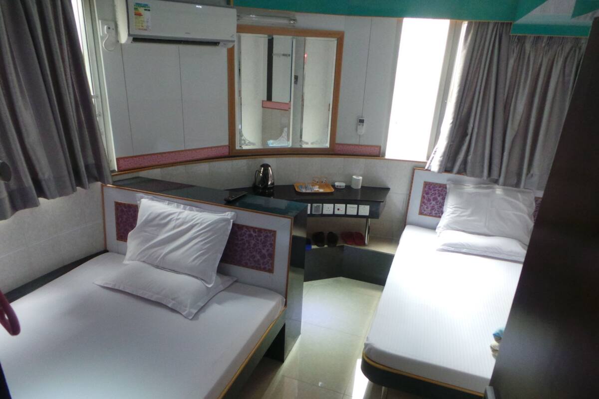 A hotel room with two single beds, a small table with a kettle, a mirror, and a shelf with toiletries.