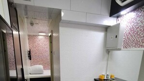 A small hotel room with a bed, a bedside table with a lamp, a small trash bin, and a bathroom visible through an open door. - Yue Ka Hotel (Kowloon)