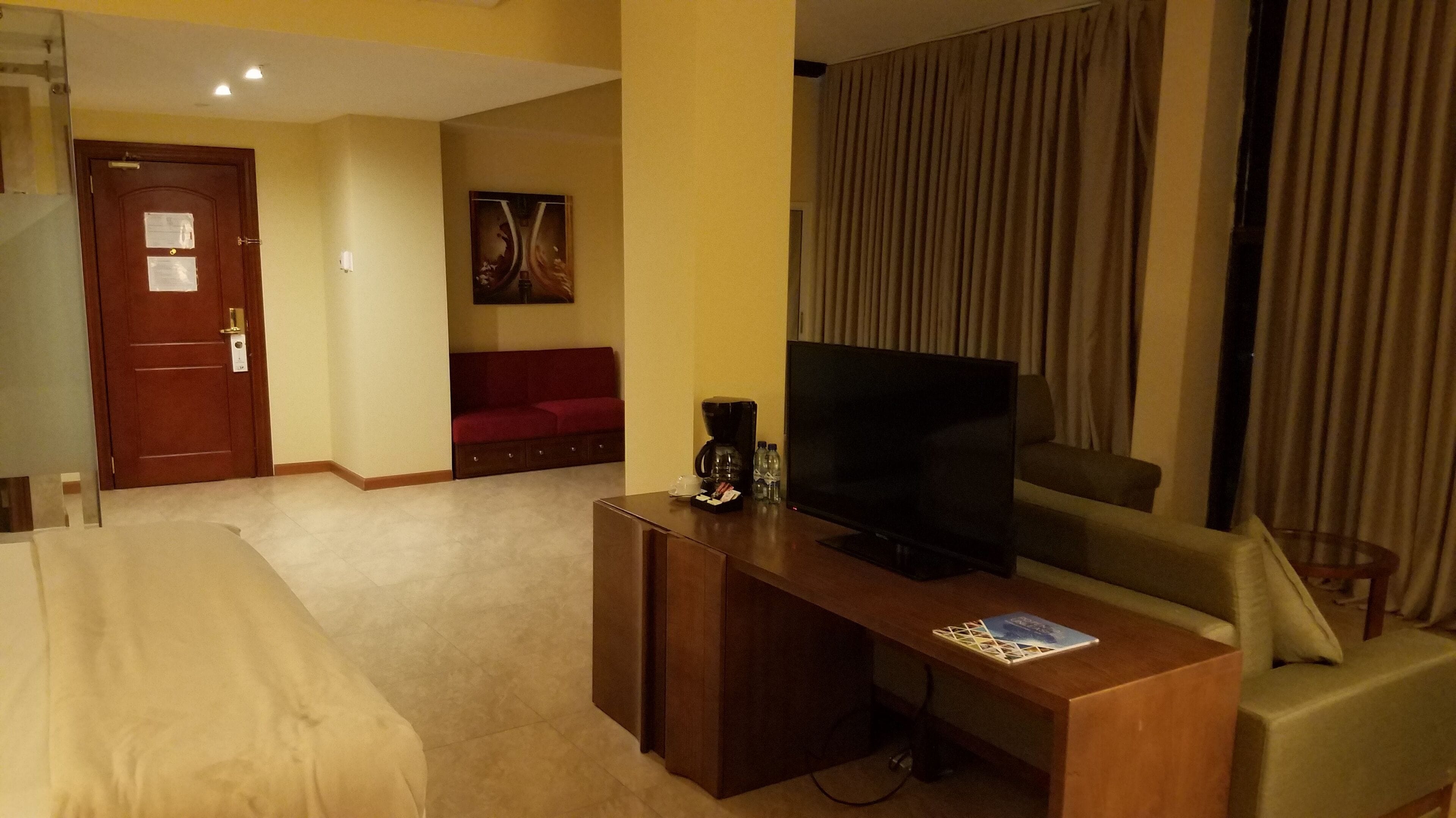 A hotel room with a bed, a TV, a coffee maker, a sofa, and a large window with curtains.