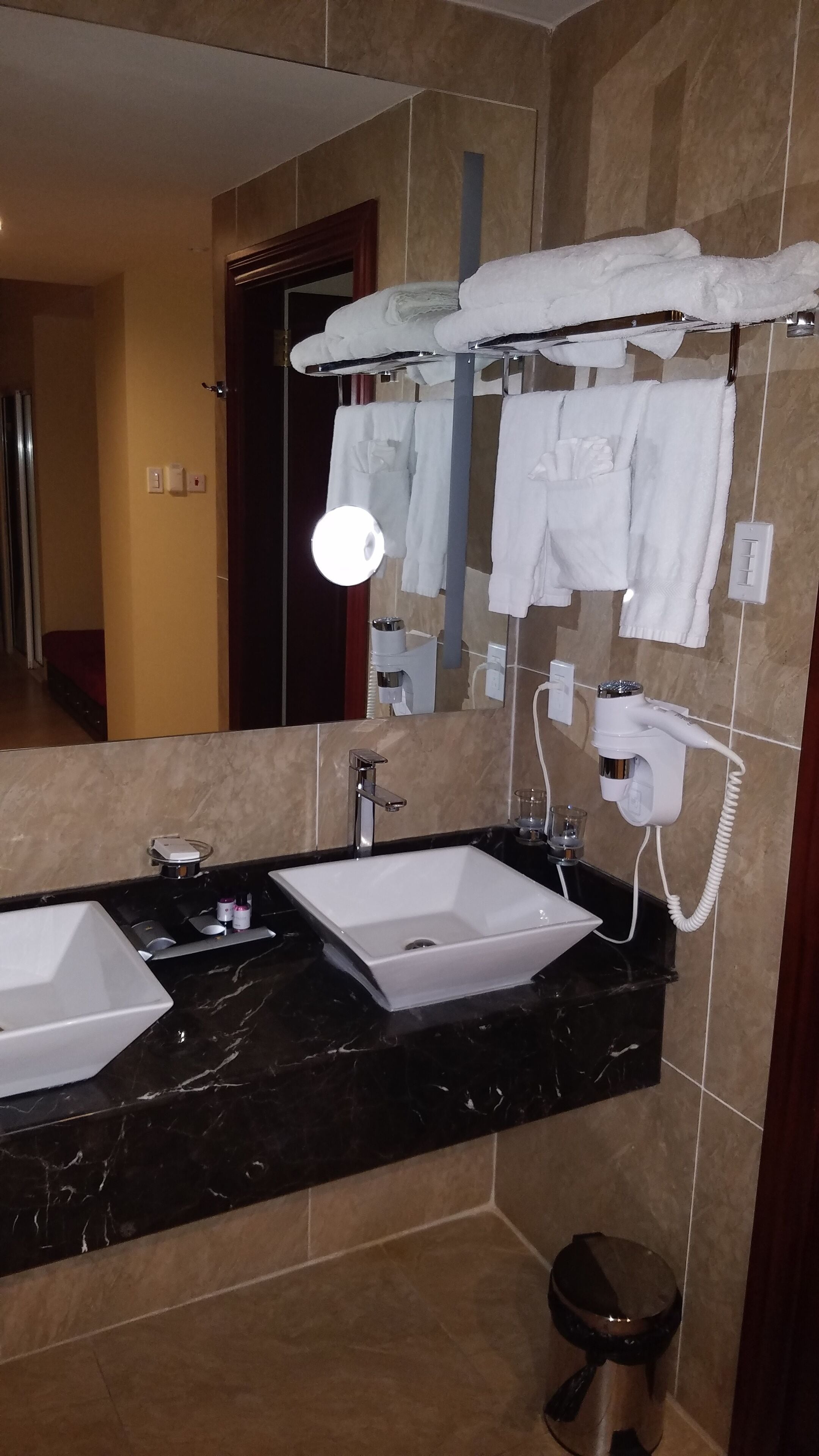 Une salle de bain avec deux lavabos, un miroir, un porte-serviettes avec des serviettes, un sèche-cheveux et une poubelle.