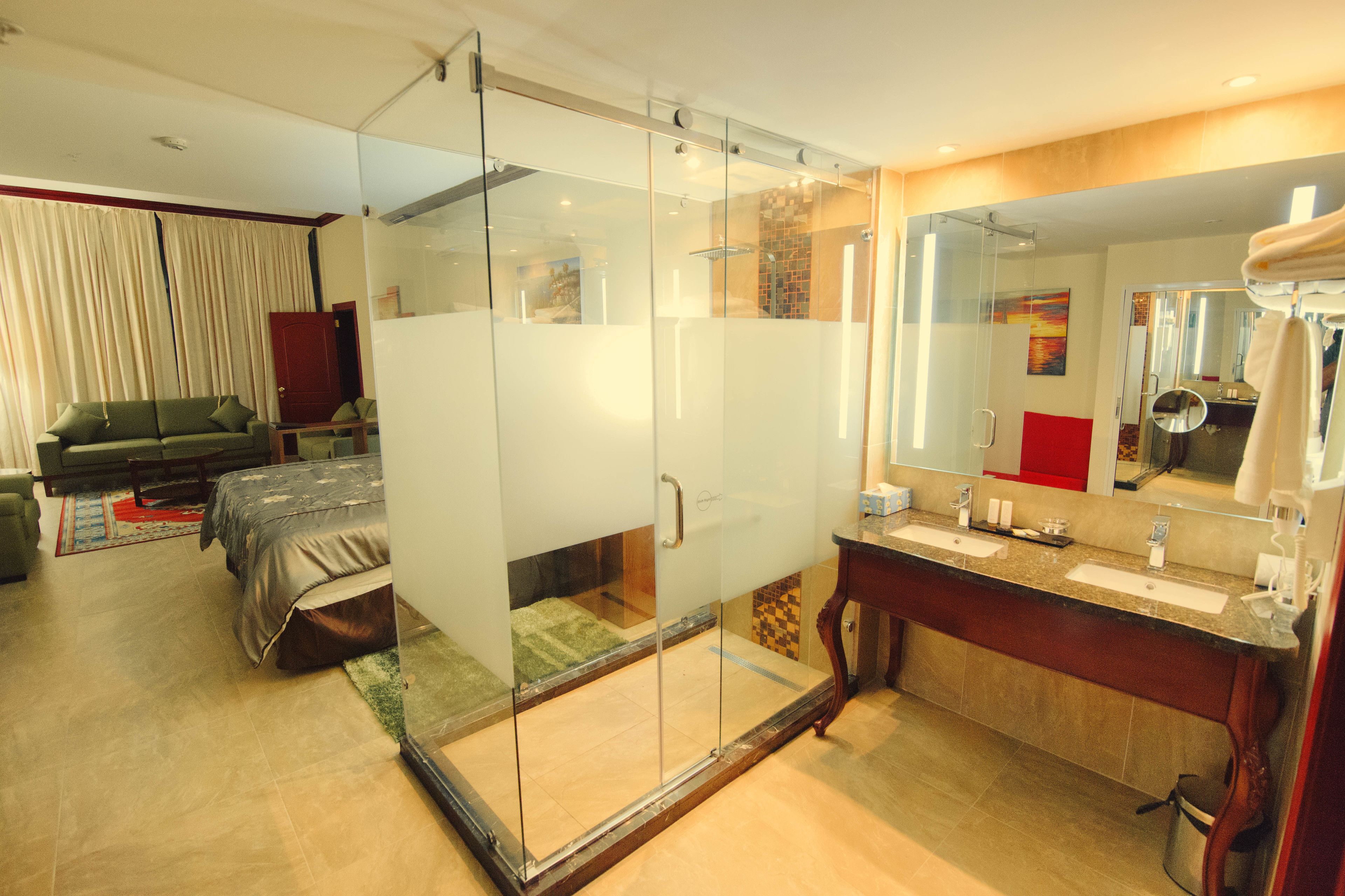 Een moderne hotelkamer met een douchecabine van glas, een apart slaapgedeelte met een bed, en een badkamer met een grote spiegel en twee wastafels.