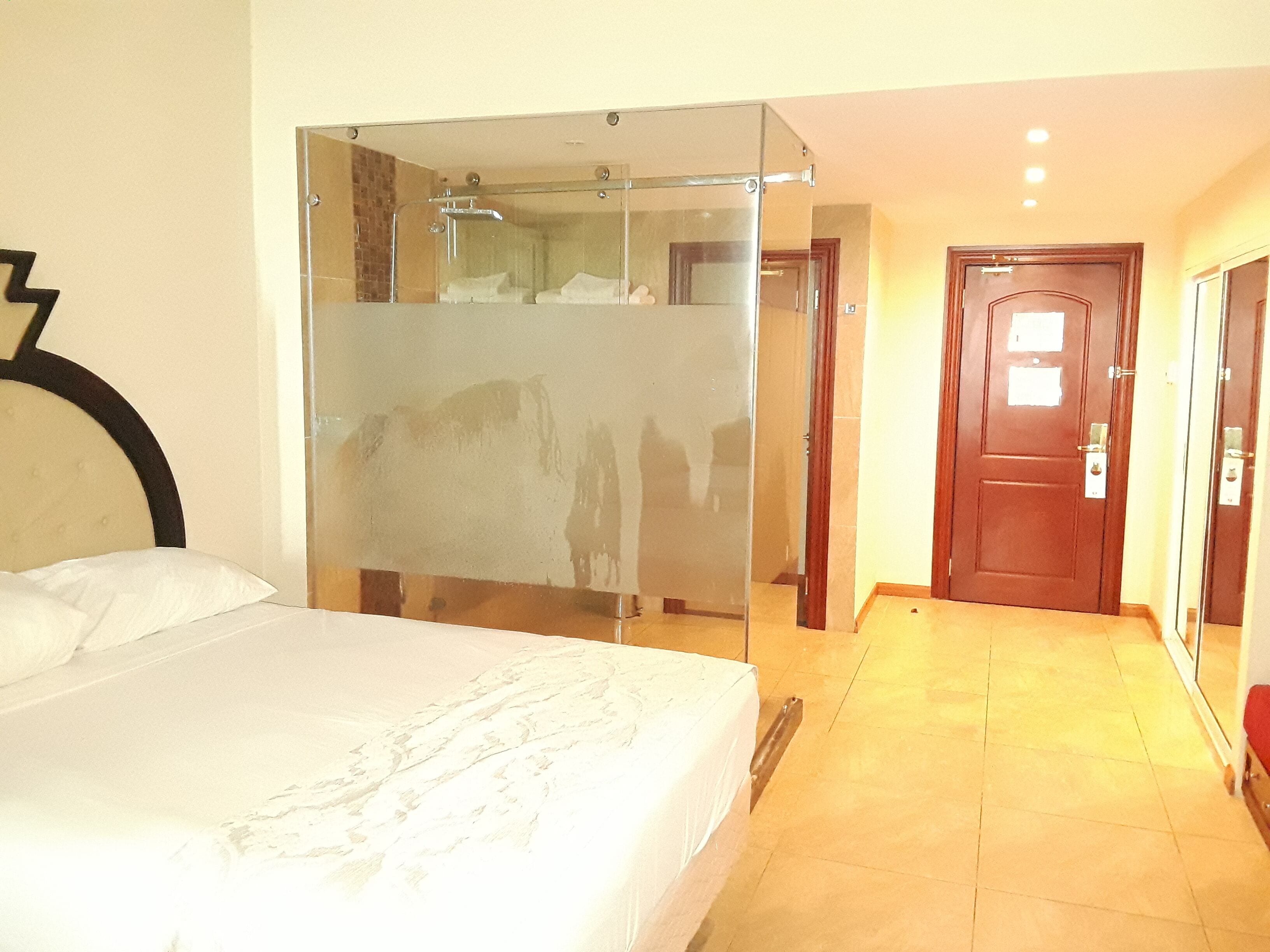 A hotel room with a bed, a glass partition, a wooden door, and a tiled floor.