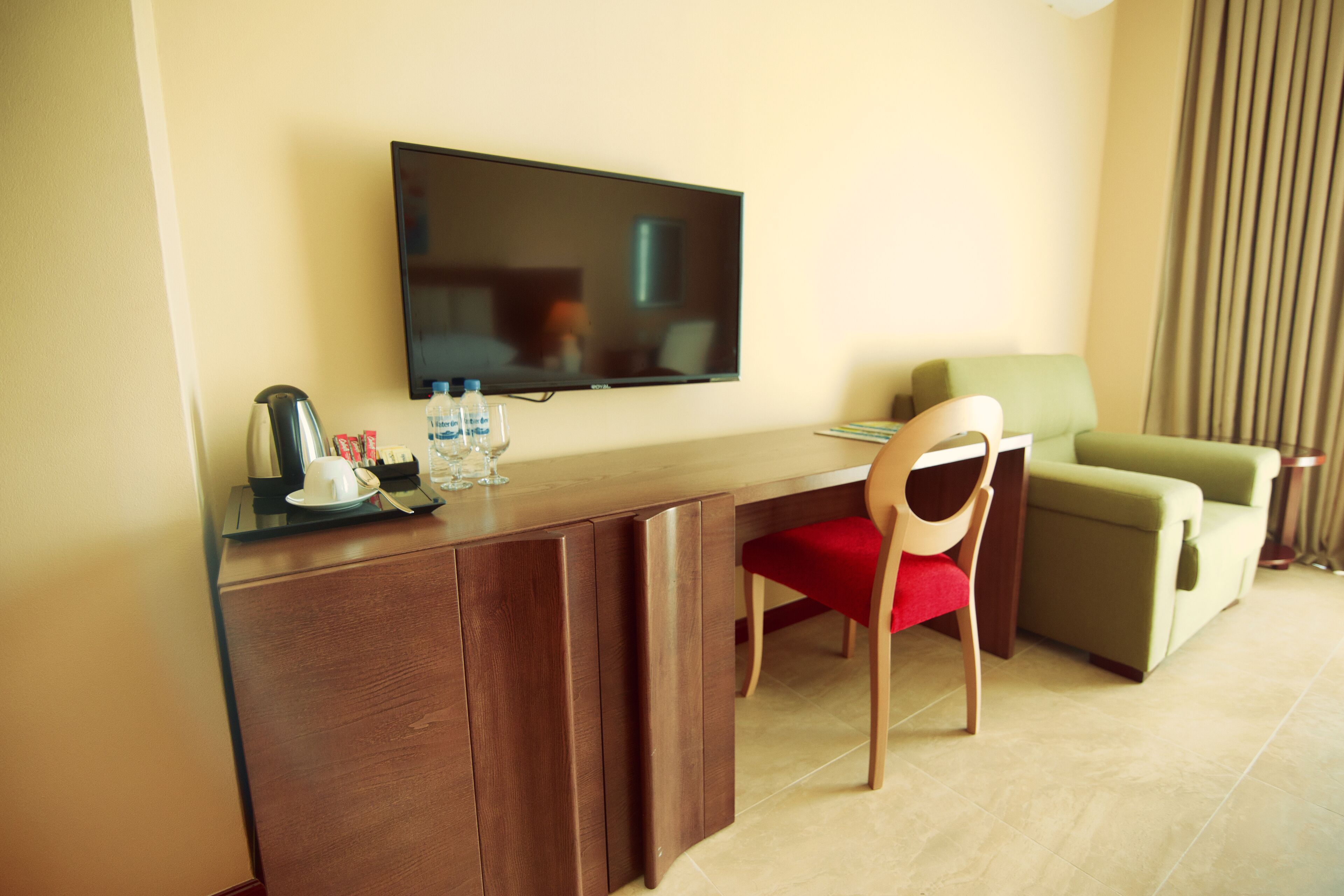 A hotel room with a wooden desk, a television mounted on the wall, a green armchair, and a sofa.