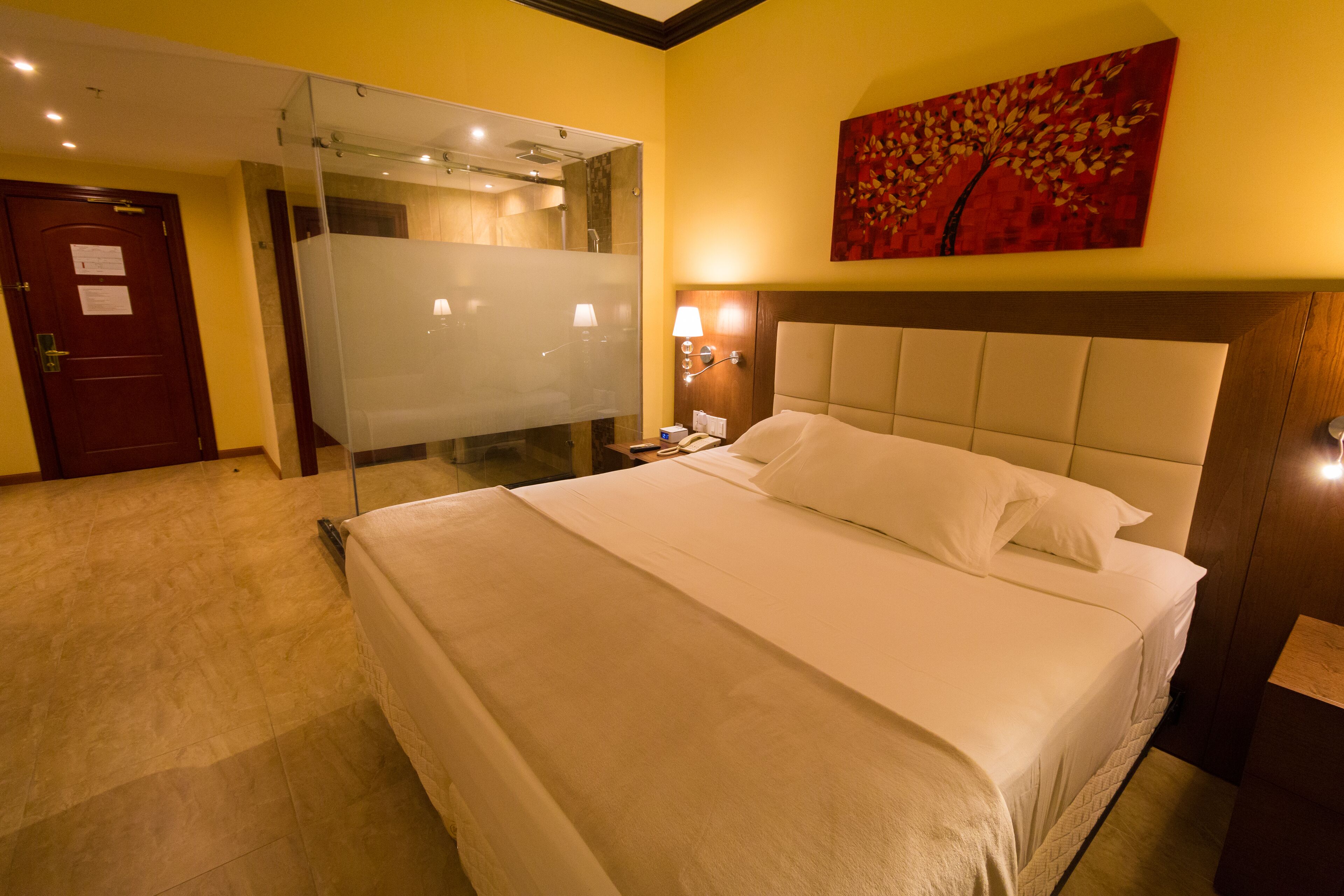 A hotel room with a large bed, a glass shower enclosure, a wooden door, and a painting on the wall.