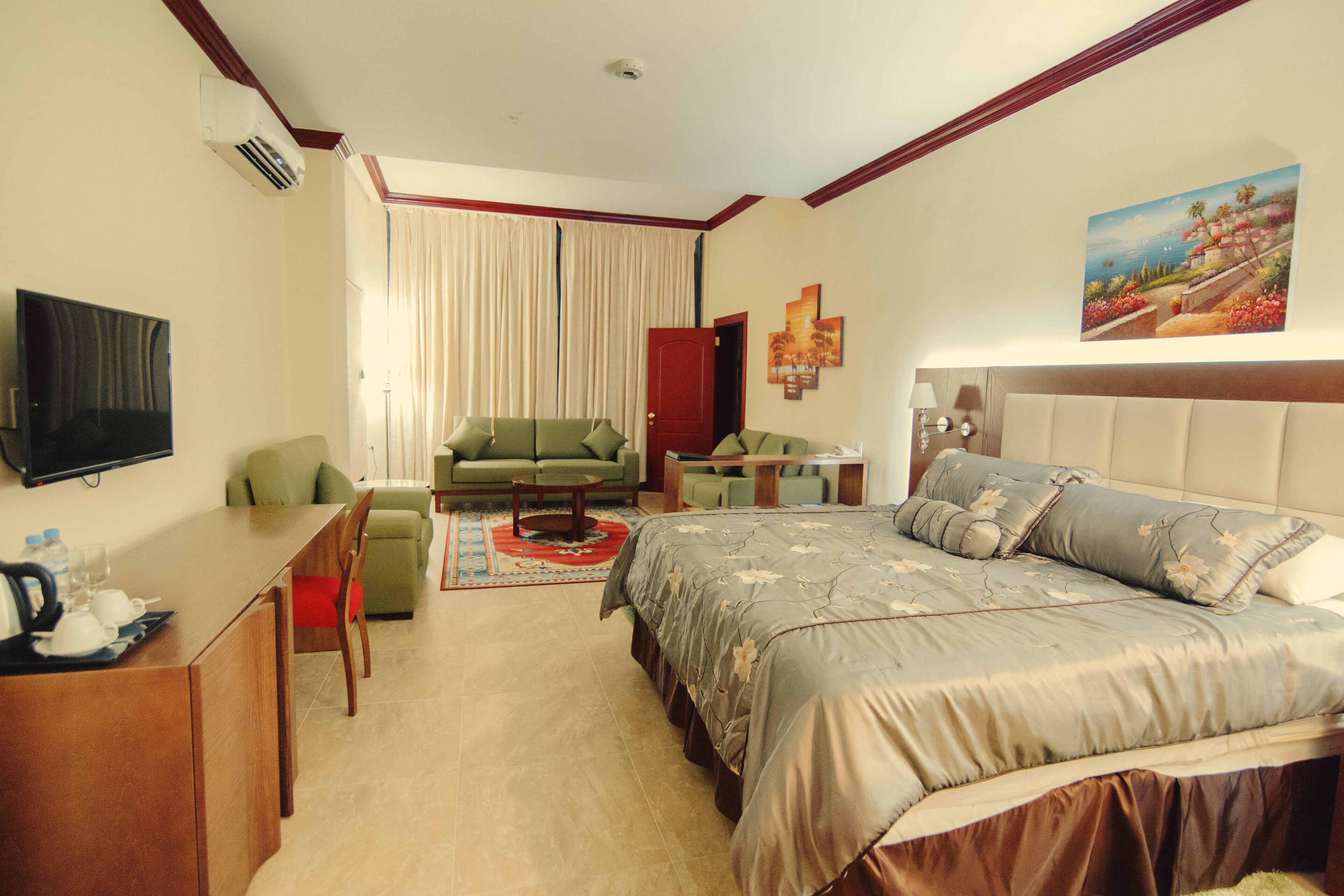 A hotel room with a bed, a sofa, a desk, a TV, and a painting on the wall.