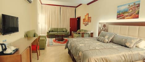 A hotel room with a bed, a sofa, a desk, a TV, and a painting on the wall.