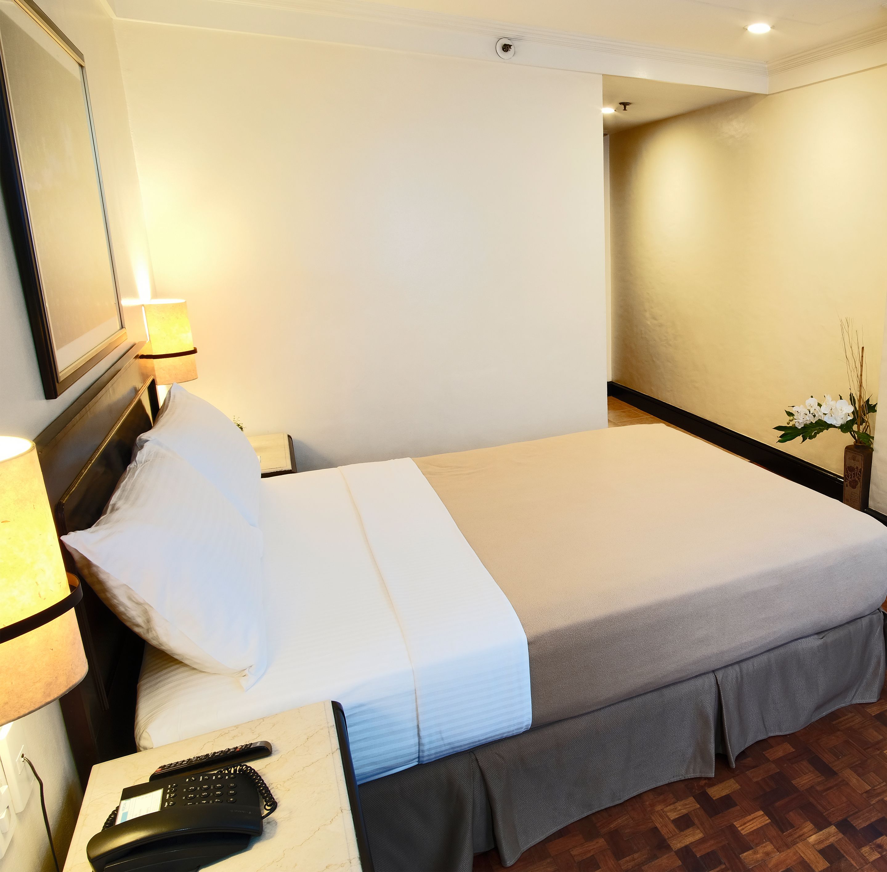 A hotel room with a bed, a nightstand with a lamp, a phone, and a vase with flowers.