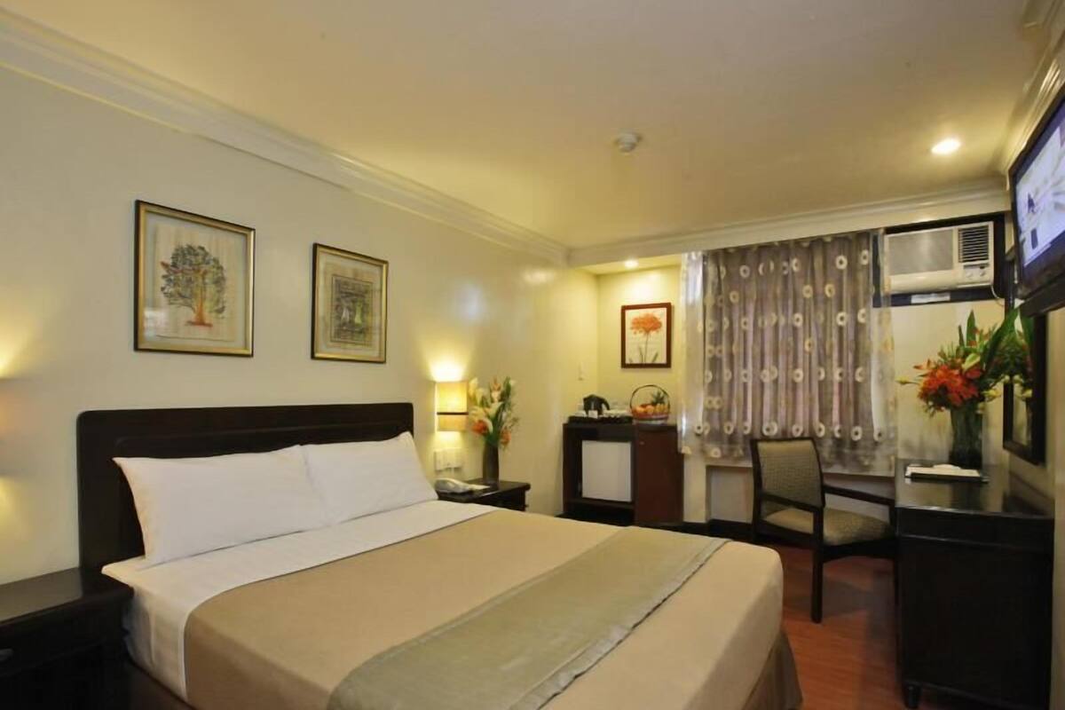 A hotel room with a bed, a desk, a chair, a nightstand, a lamp, and framed pictures on the wall.