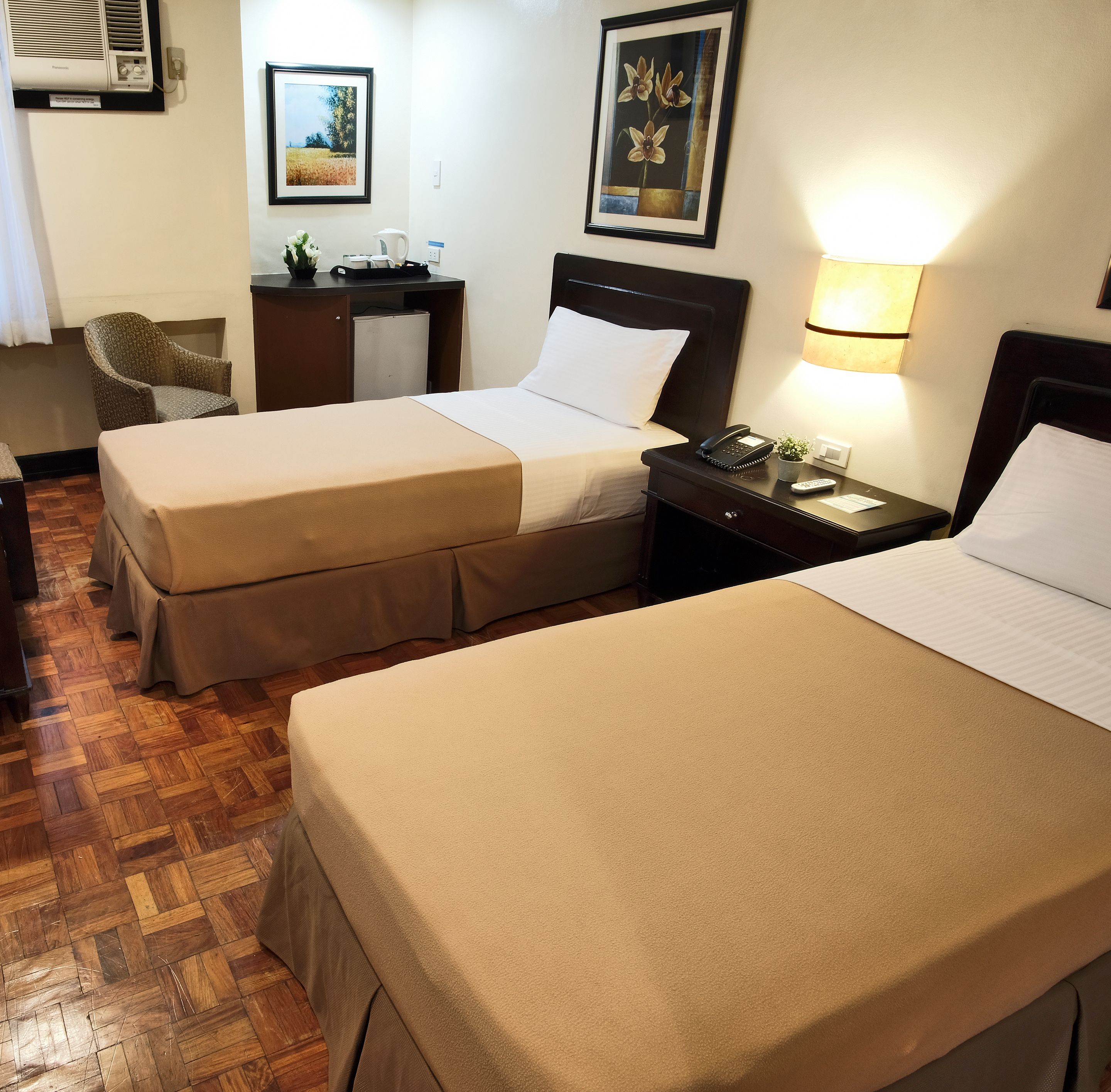 A hotel room with two beds, a desk with a computer, a chair, and a wall-mounted air conditioner.