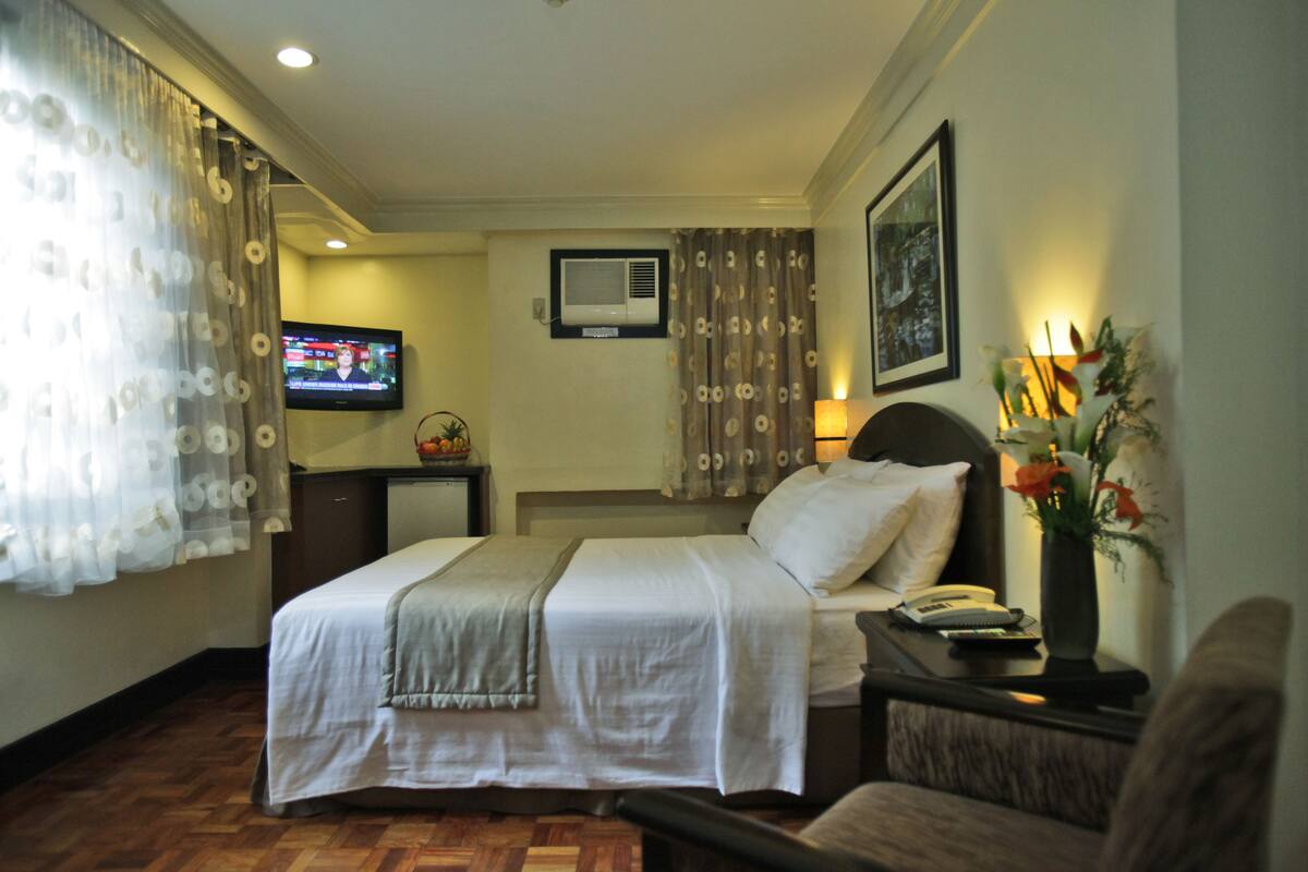 A hotel room with a bed, a desk with a lamp, a TV, and a wall-mounted air conditioner.