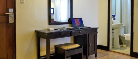 Una habitación de hotel con un escritorio de madera, una computadora portátil, un banco y un jarrón de flores.