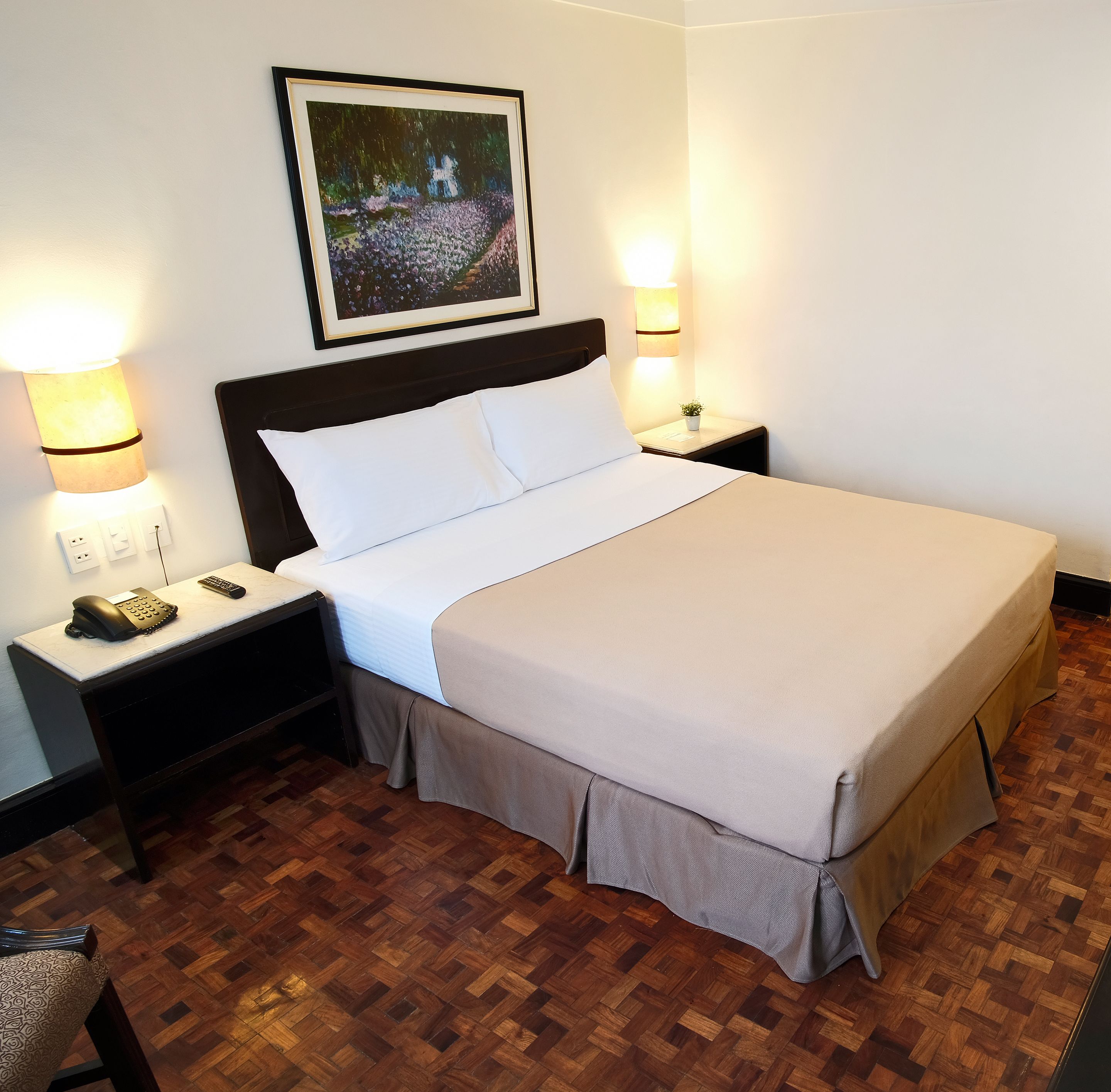 A neatly made bed with white linens, a dark headboard, and a small bedside table with a lamp and a phone. The room has a wooden parquet floor and a framed picture on the wall.