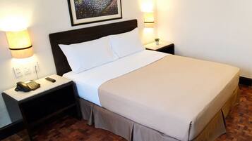 A neatly made bed with white linens, a dark headboard, and a small bedside table with a lamp and a phone. The room has a wooden parquet floor and a framed picture on the wall.