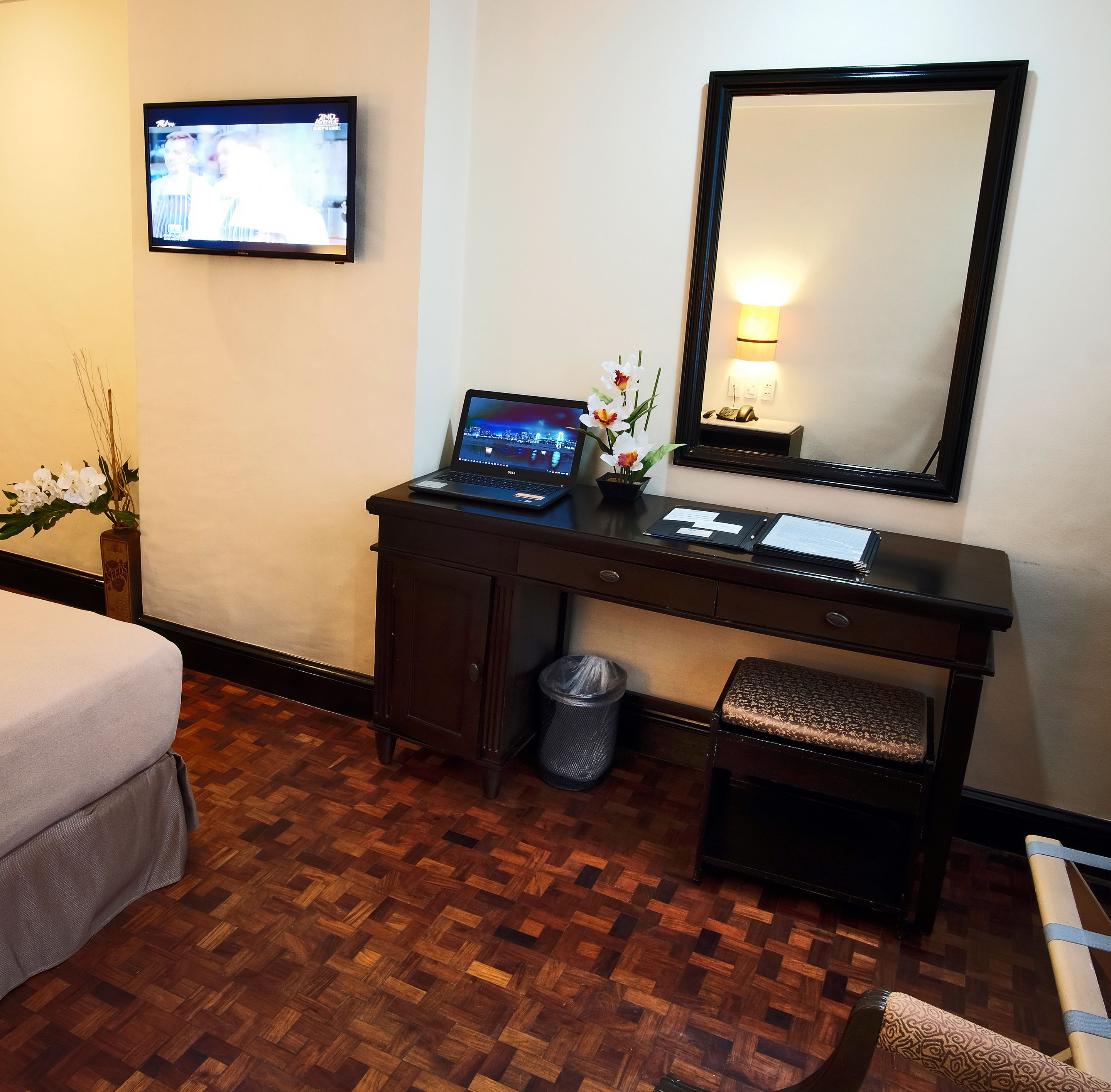 Hotelkamer met een houten bureau, een laptop, een kleine plant in pot, een prullenbak, een bankje, een bed, een televisie en een spiegel aan de muur.