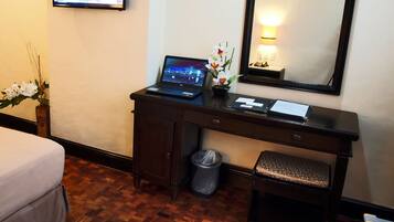 Hotelkamer met een houten bureau, een laptop, een kleine plant in pot, een prullenbak, een bankje, een bed, een televisie en een spiegel aan de muur.