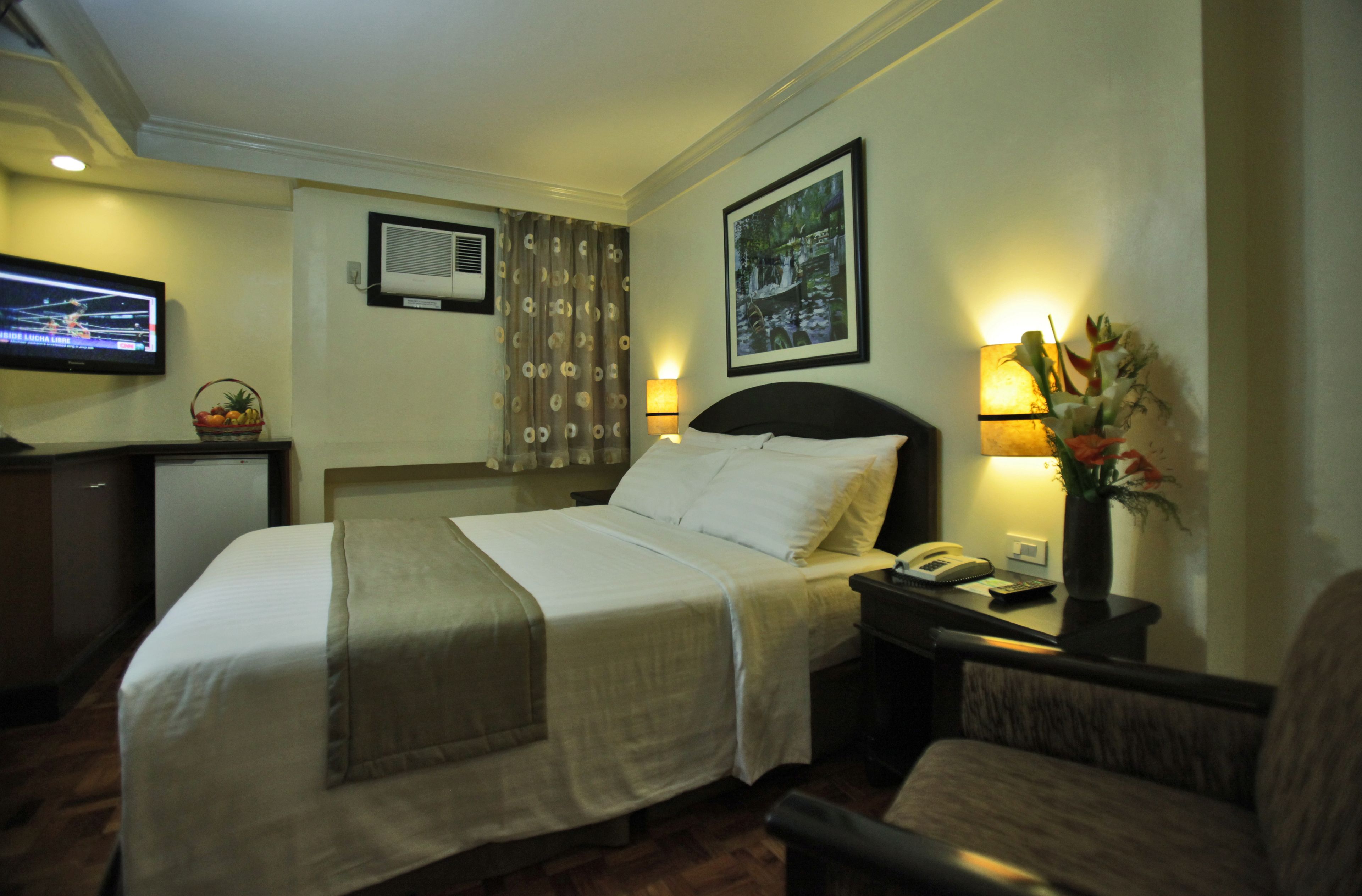 A hotel room with a bed, a desk with a lamp, a television, and a wall-mounted air conditioner.