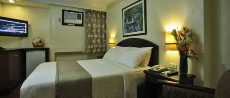 A hotel room with a bed, a desk with a lamp, a television, and a wall-mounted air conditioner.