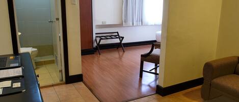 A hotel room with a wooden floor, a desk with paperwork, a chair, a sofa, a bed, a window with curtains, and an air conditioning unit.