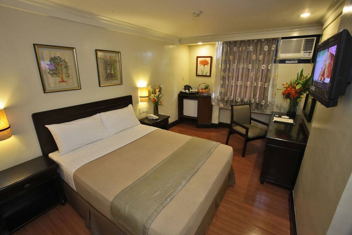 A hotel room with a bed, bedside table, lamp, desk, chair, TV, and artwork on the walls.