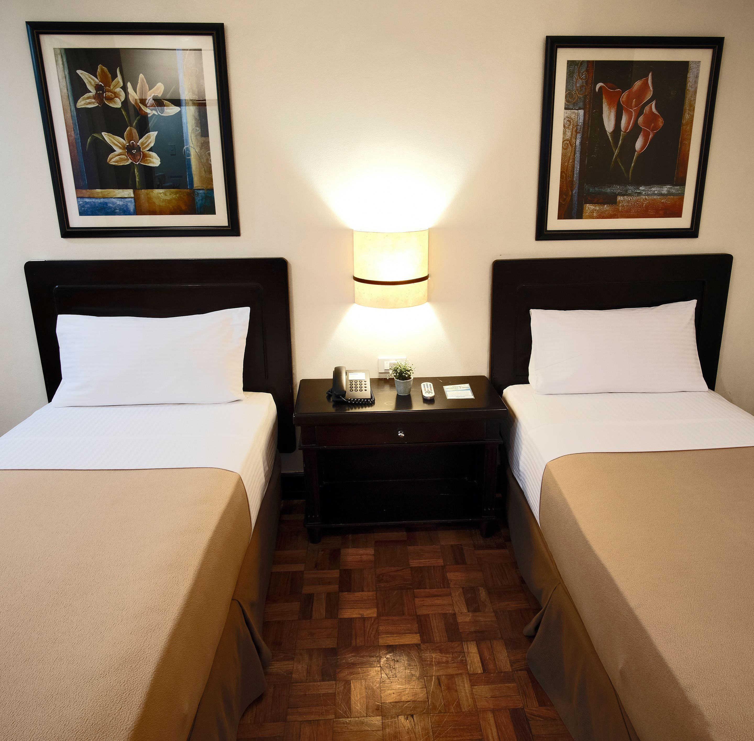 A hotel room with two beds, a nightstand with a lamp, and two framed floral paintings on the wall.