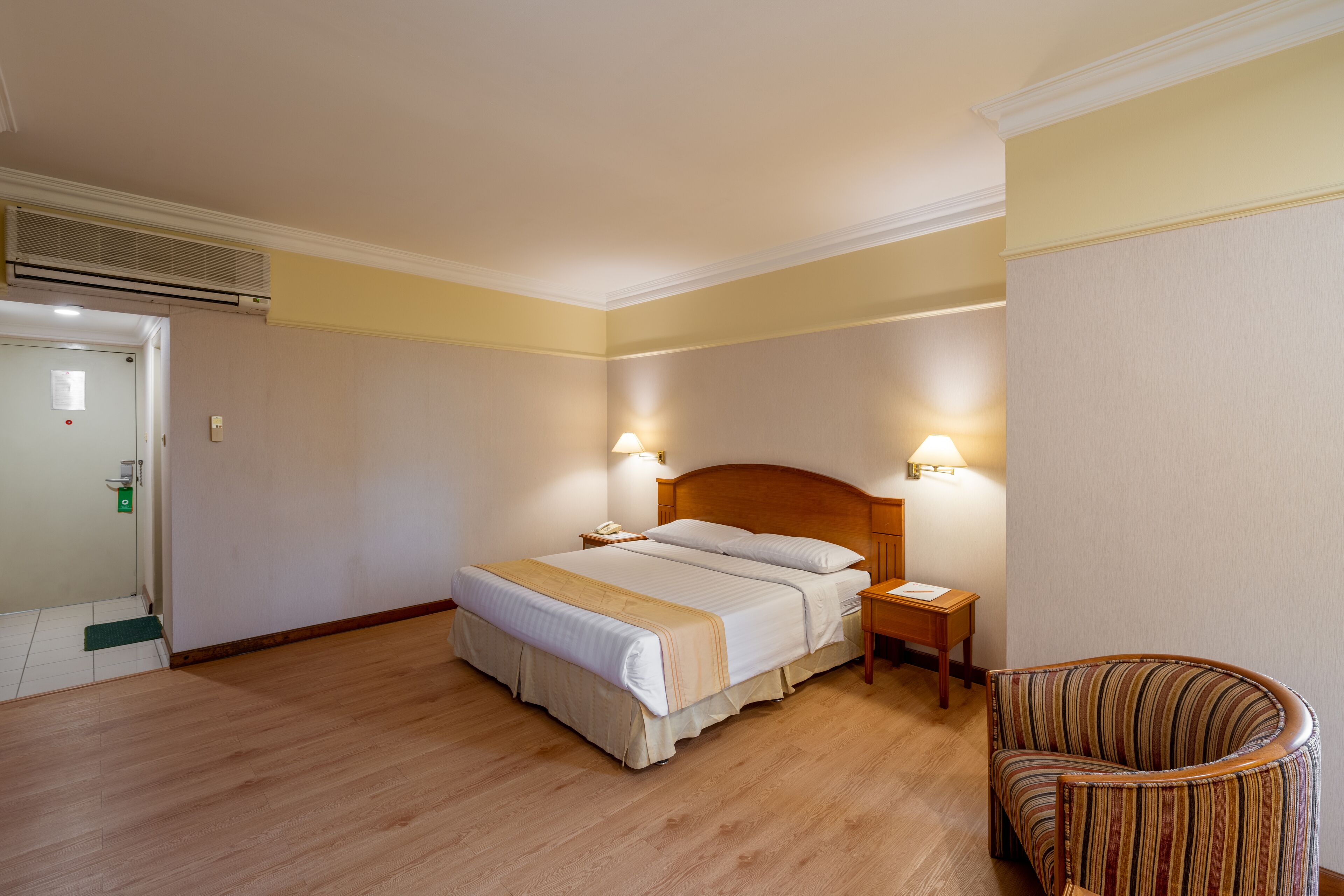 Quarto de hotel com piso de madeira, uma cama de casal com encosto, duas luminárias de cabeceira, uma cadeira e uma mesinha.