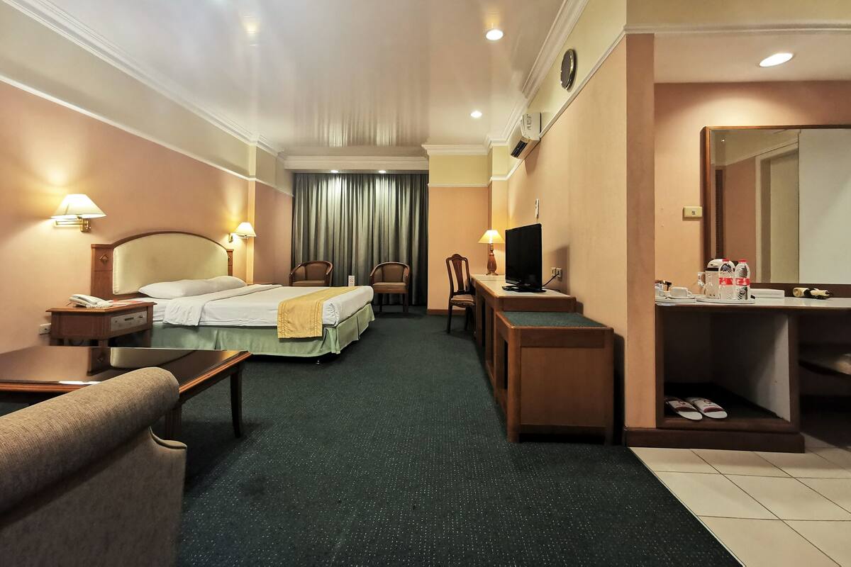 Habitación de hotel con cama, dos sillas, escritorio con televisor, espejo y zapatero.