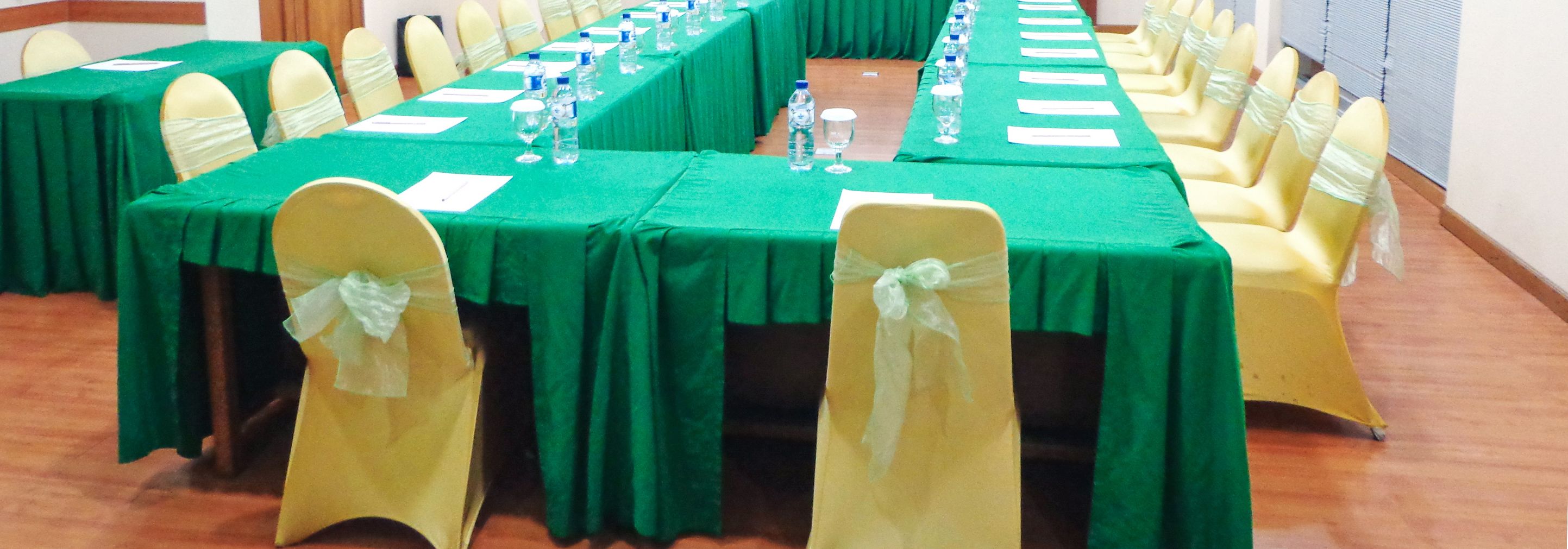 A conference room with green tables and yellow chairs, equipped with water bottles and glasses.