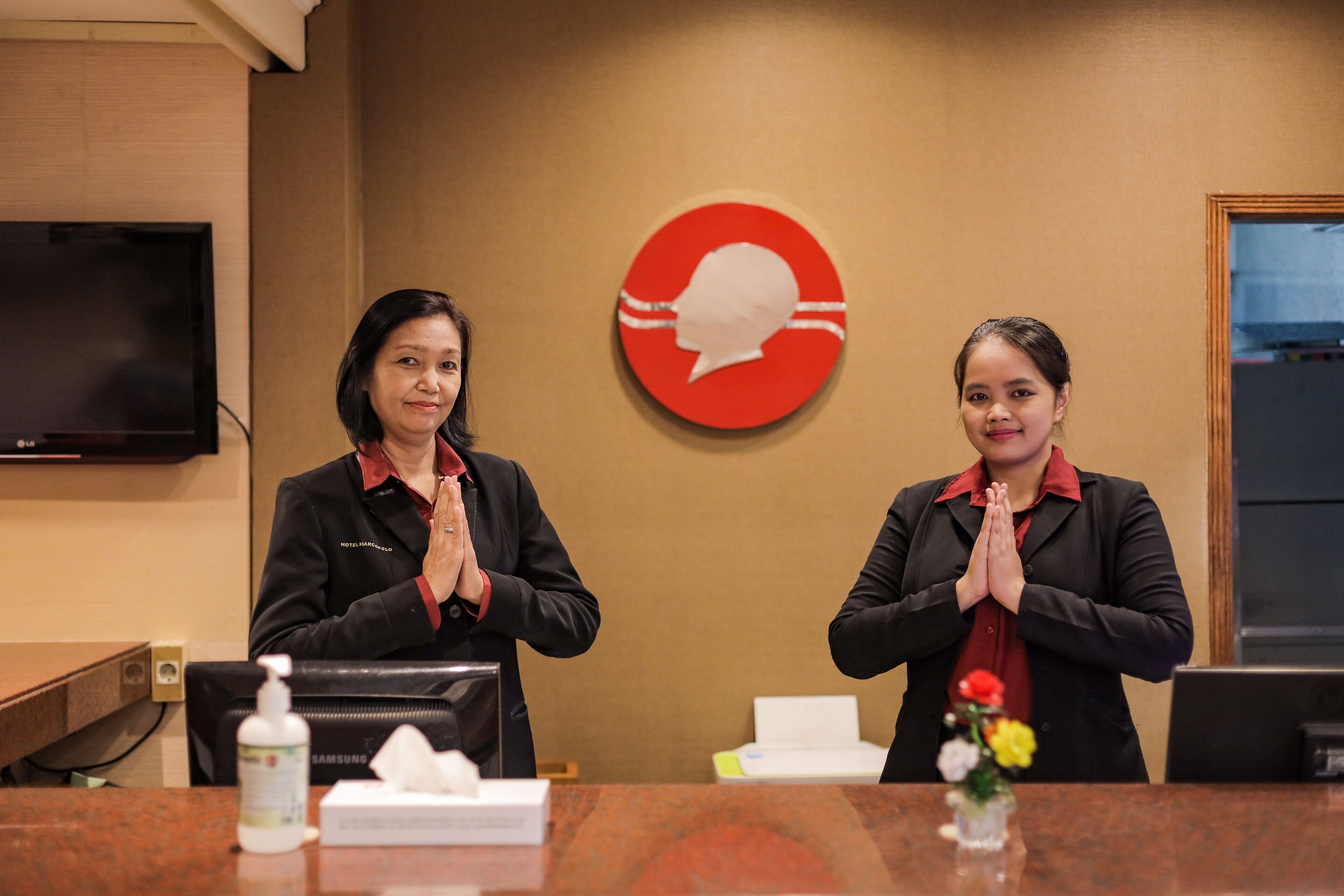 Two receptionists in a hotel lobby with a television on the wall, a red circular sign, and a flower vase on the counter.