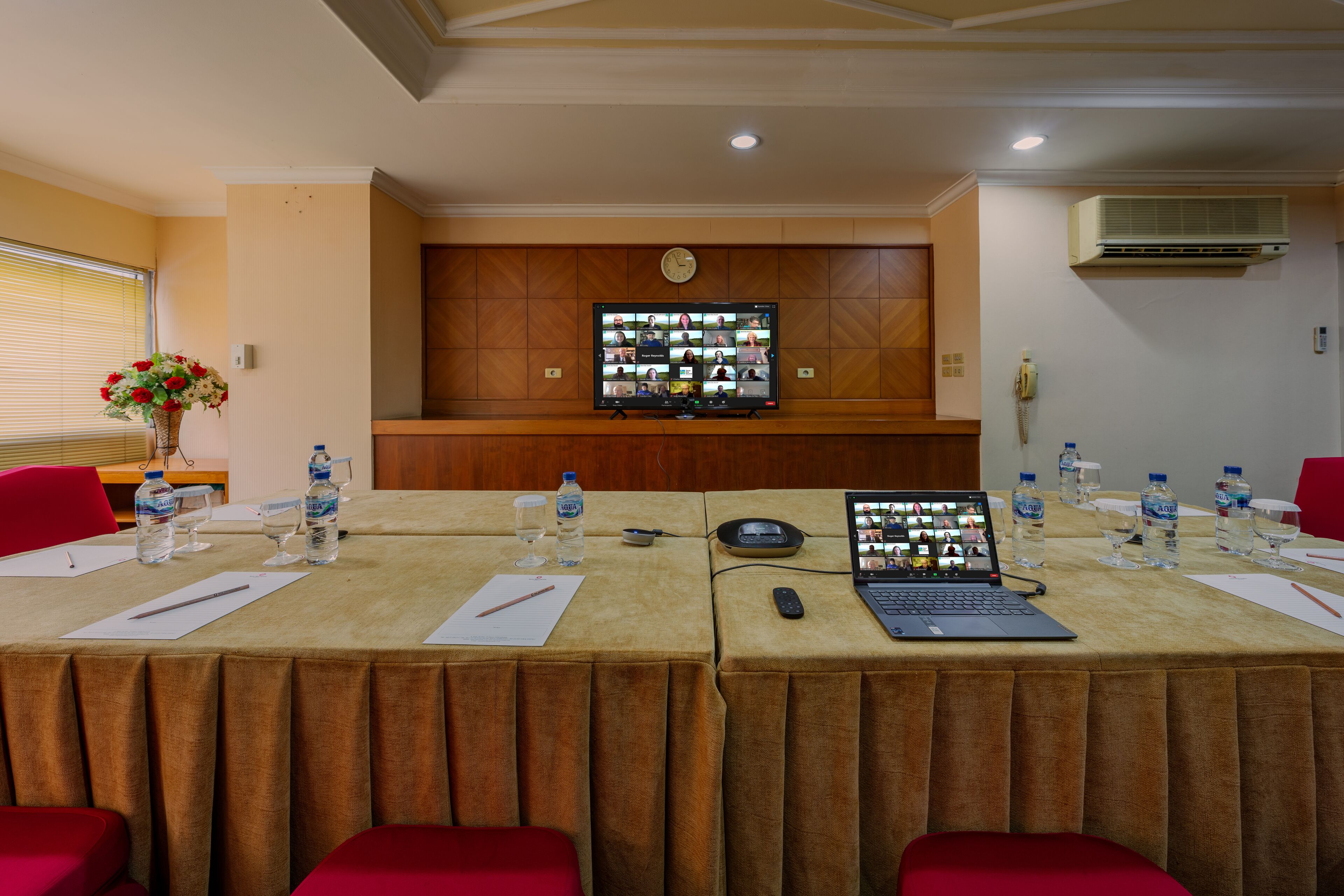 A conference room with a long table, red chairs, a laptop, and a large screen displaying multiple video feeds.
