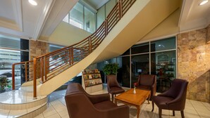 A lobby with a curved staircase, leather seating, a wooden coffee table, and a reception area with a plant and magazine rack. - Hotel Marcopolo Jakarta (Jakarta)