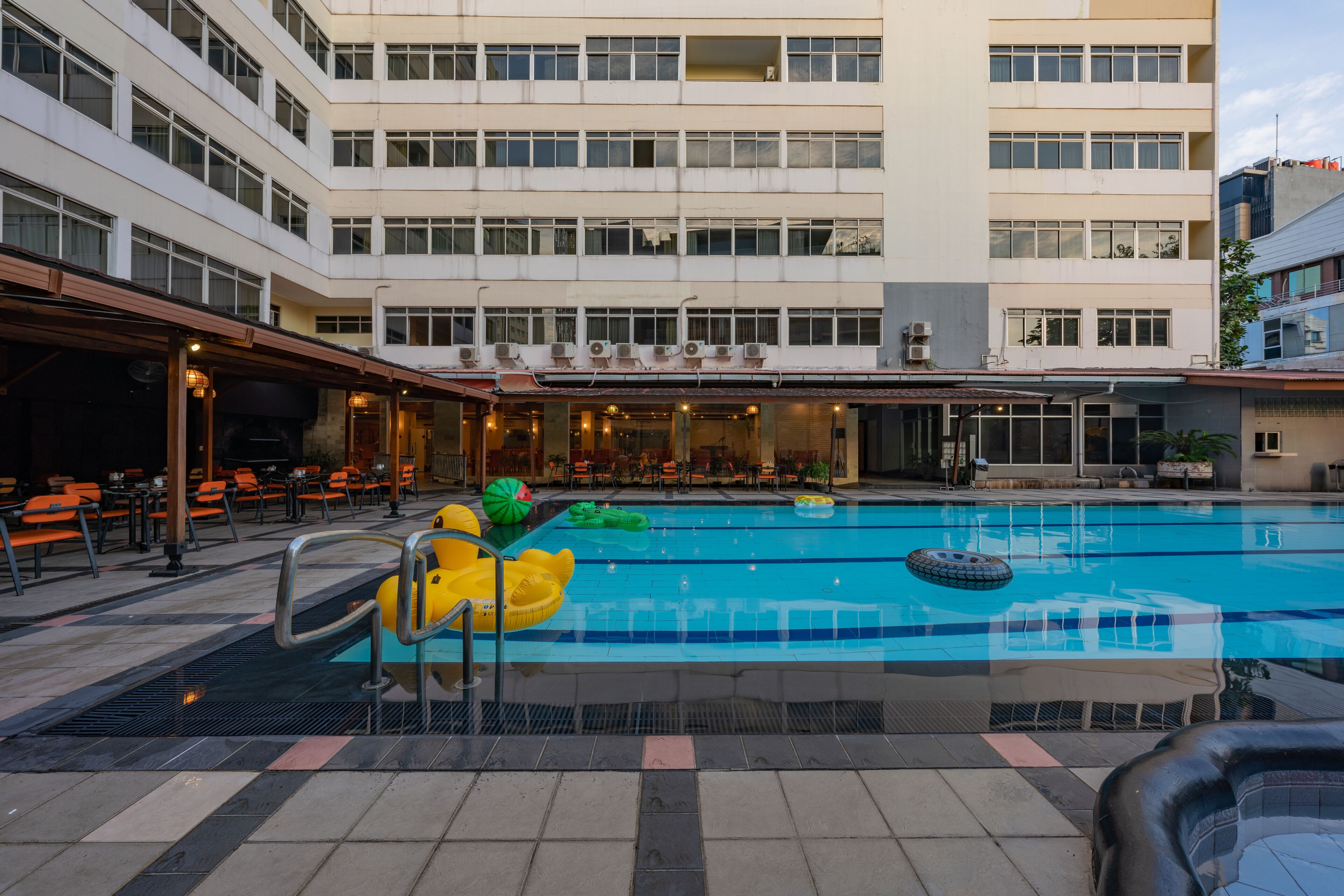 Une piscine entourée de chaises longues et de parasols, elle-même bordée d’un immeuble à plusieurs étages doté de balcons.