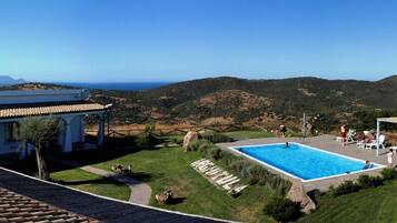 A pool area with a view of the sea and hills, surrounded by a white building with a tiled roof.