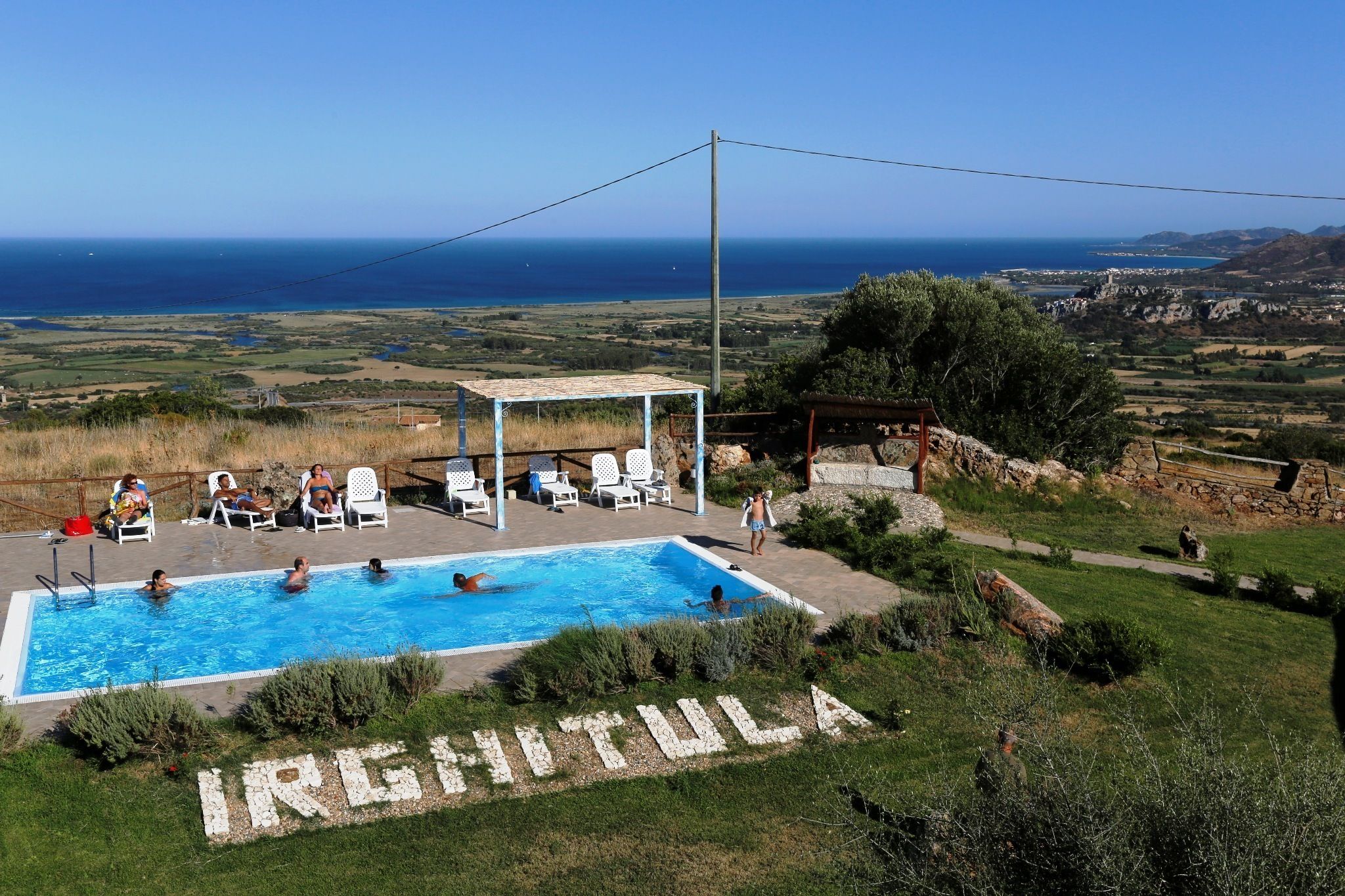 A pool area with a view of the sea, surrounded by greenery and a stone wall.