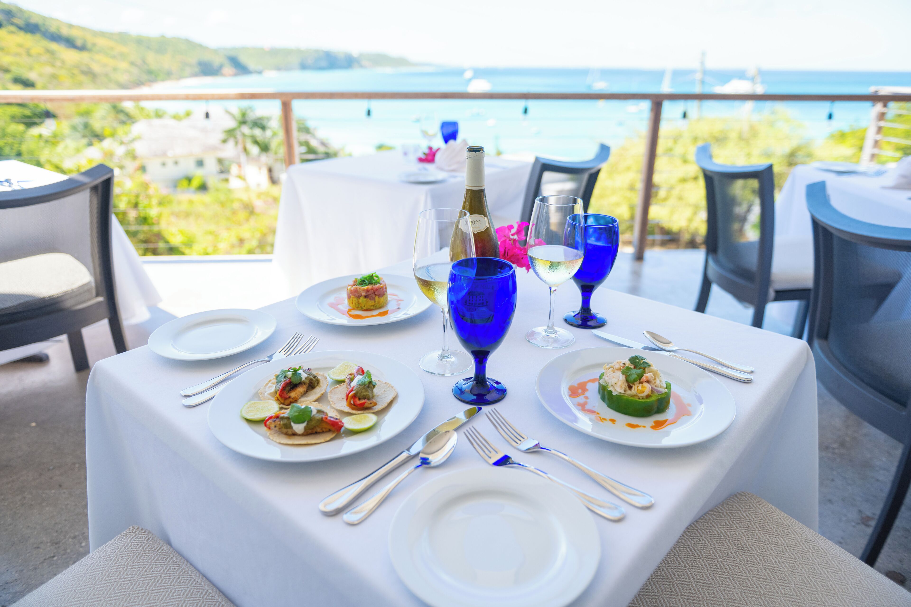 A table set for a meal with plates of food, wine glasses, and cutlery, overlooking a scenic view of the sea and hills.