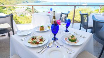 A table set for a meal with plates of food, wine glasses, and cutlery, overlooking a scenic view of the sea and hills.