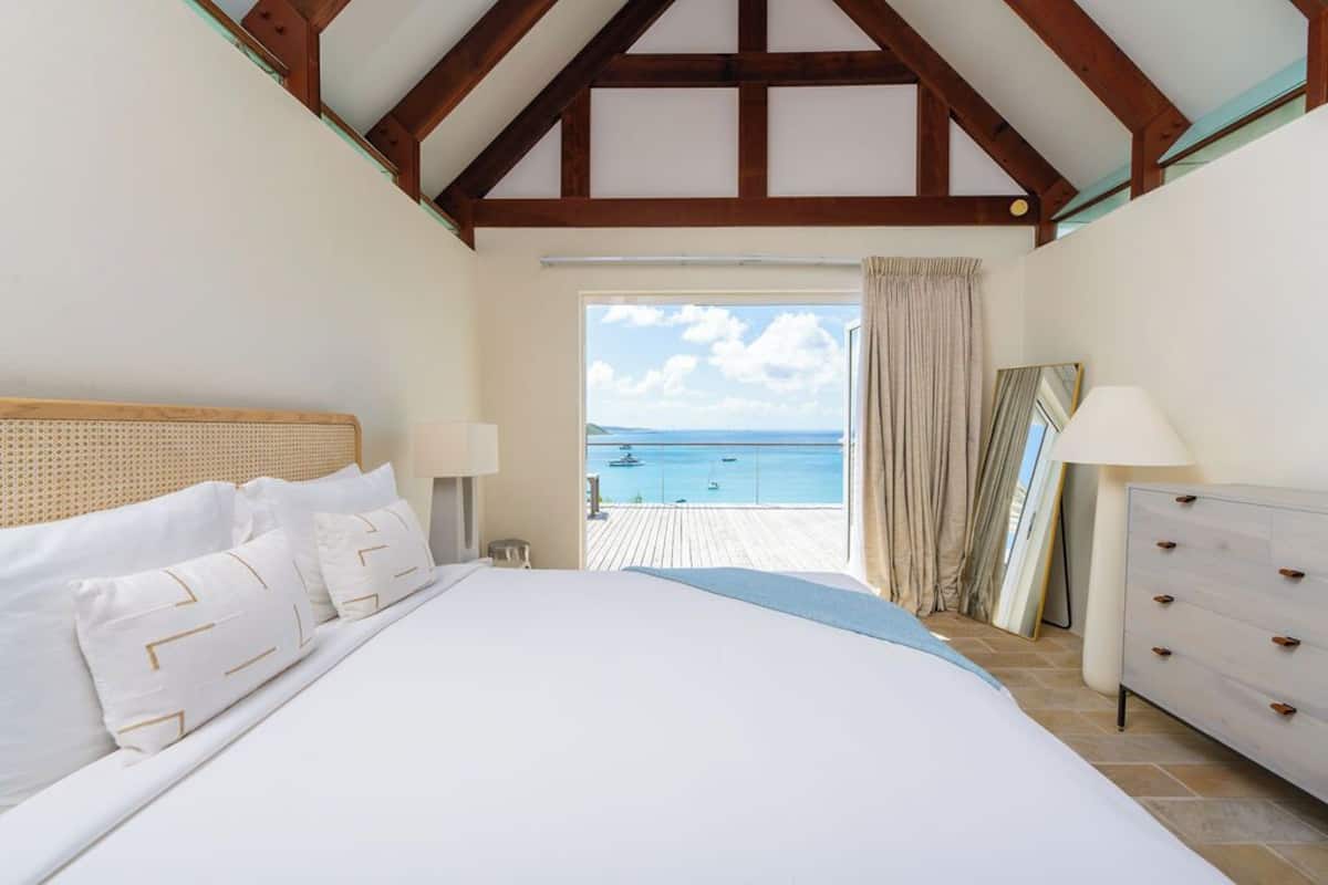 A bedroom with a large bed, a wooden headboard, a dresser, and a view of the sea through a balcony.