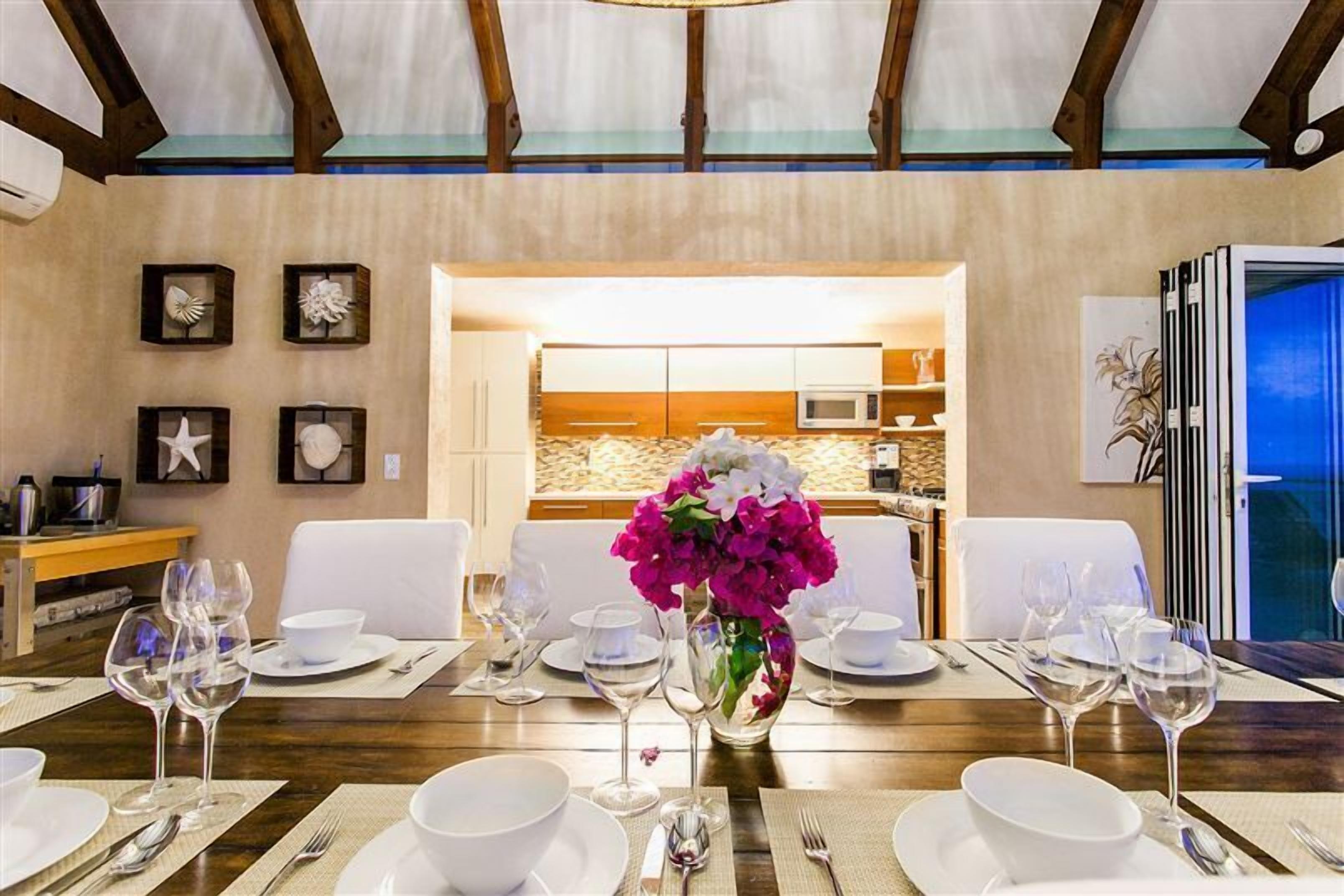 Un comedor con una mesa de madera dispuesta para seis personas, sillas blancas y un jarrón de flores.