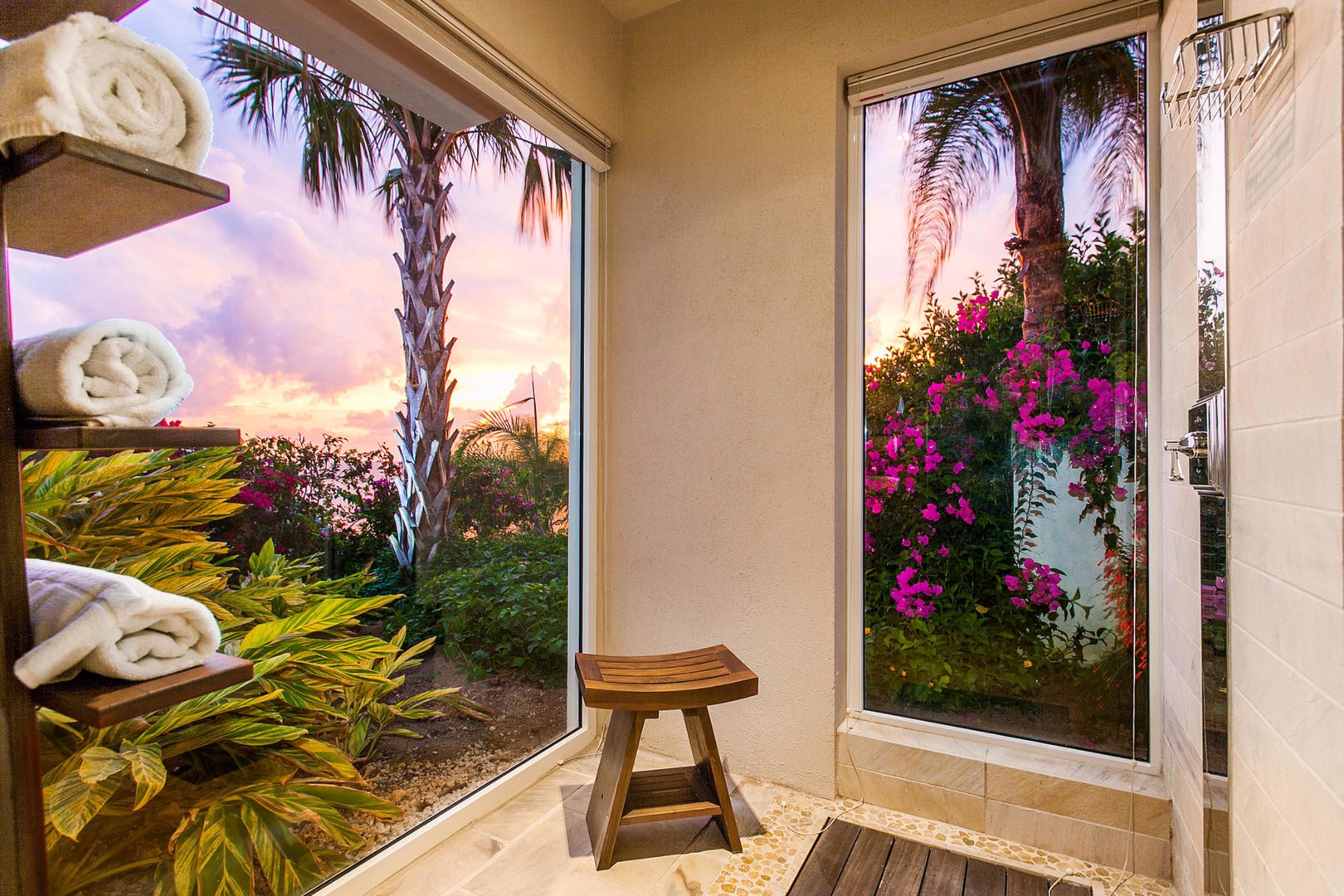 Casa de banho com um banquinho de madeira, porta-toalhas e vista para um jardim com flores cor-de-rosa e palmeiras.